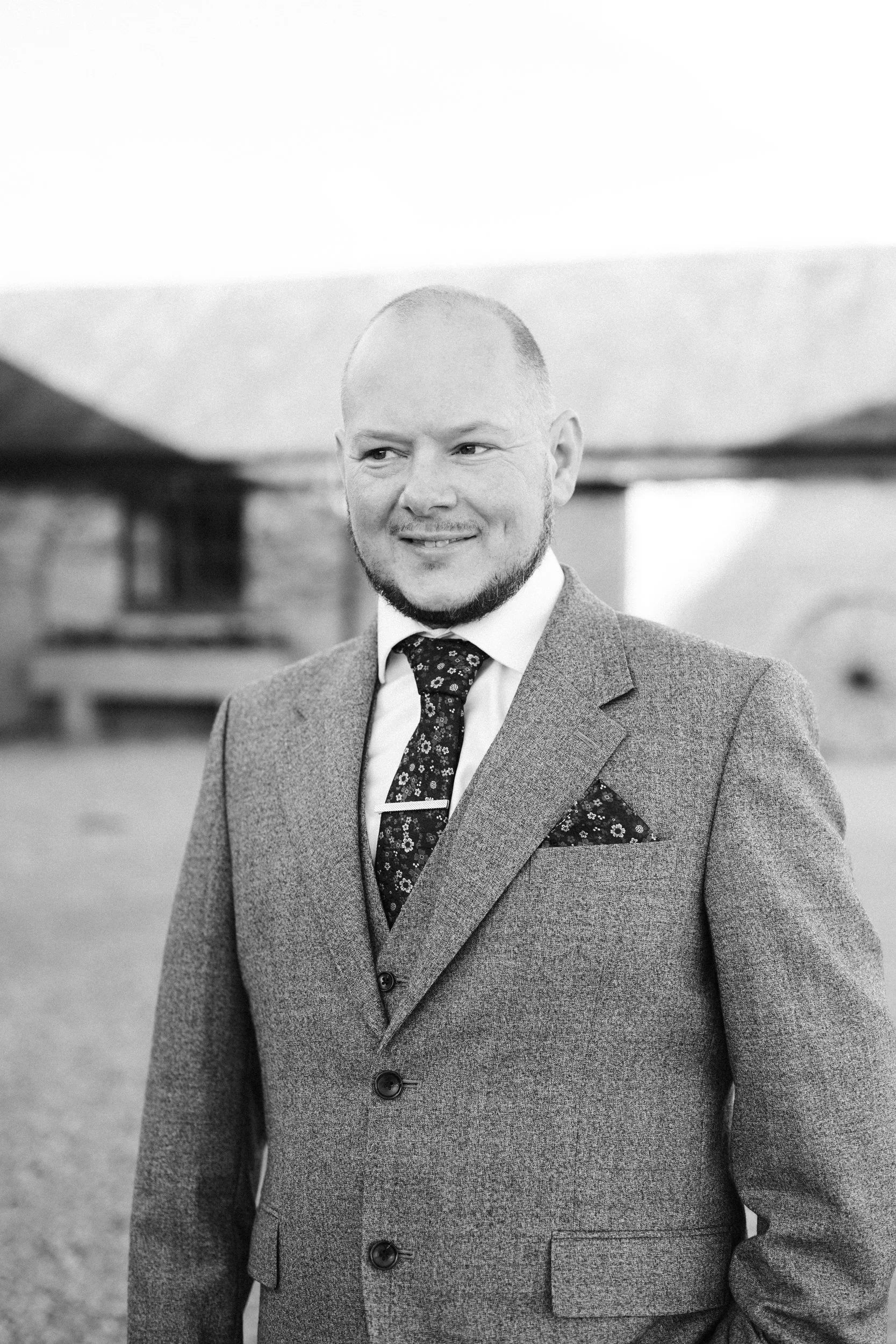 A man in a suit and tie smiling outdoors in black and white.