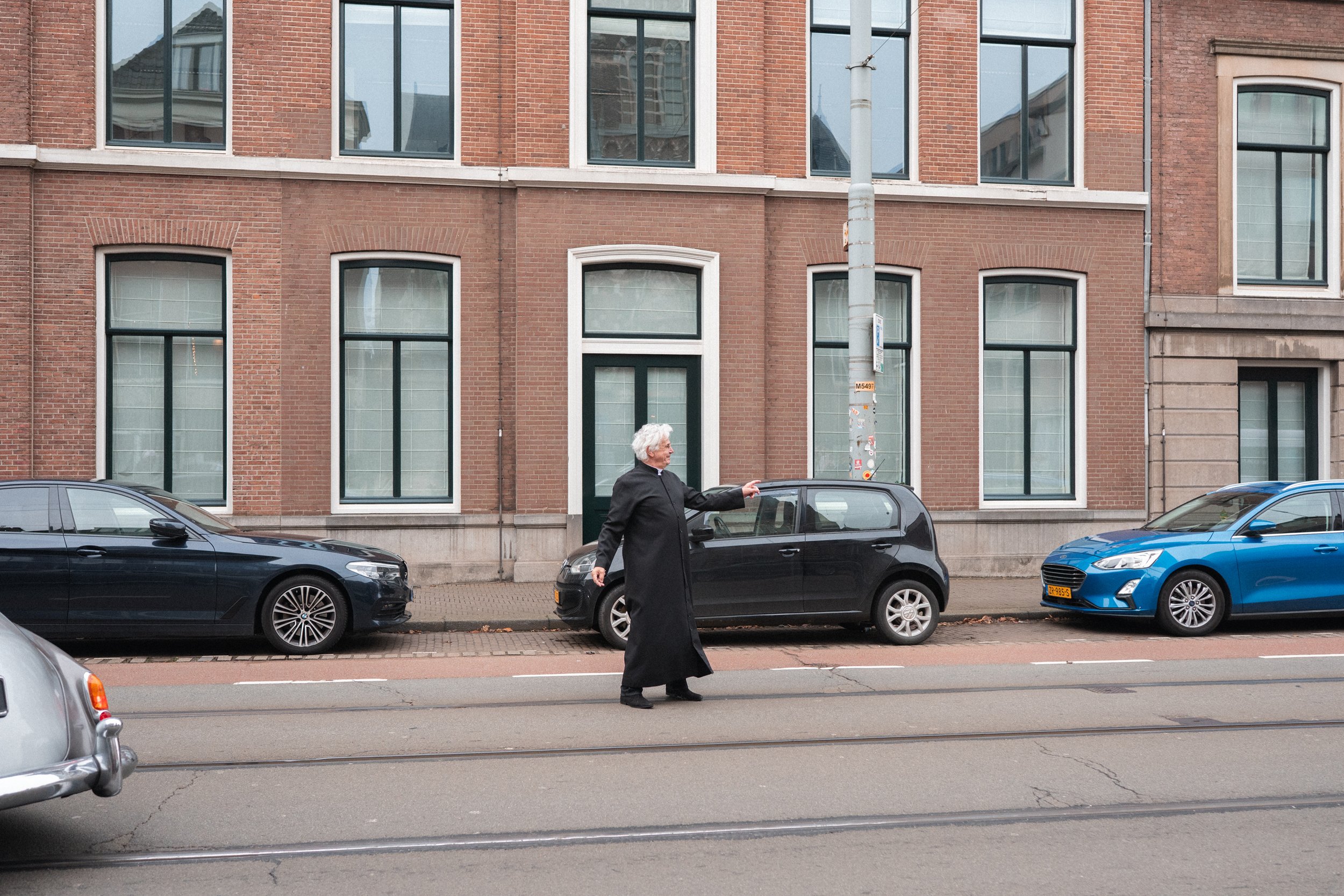 An elderly woman with white hair, wearing a long black coat, crosses the street in front of parked cars and a brick building.