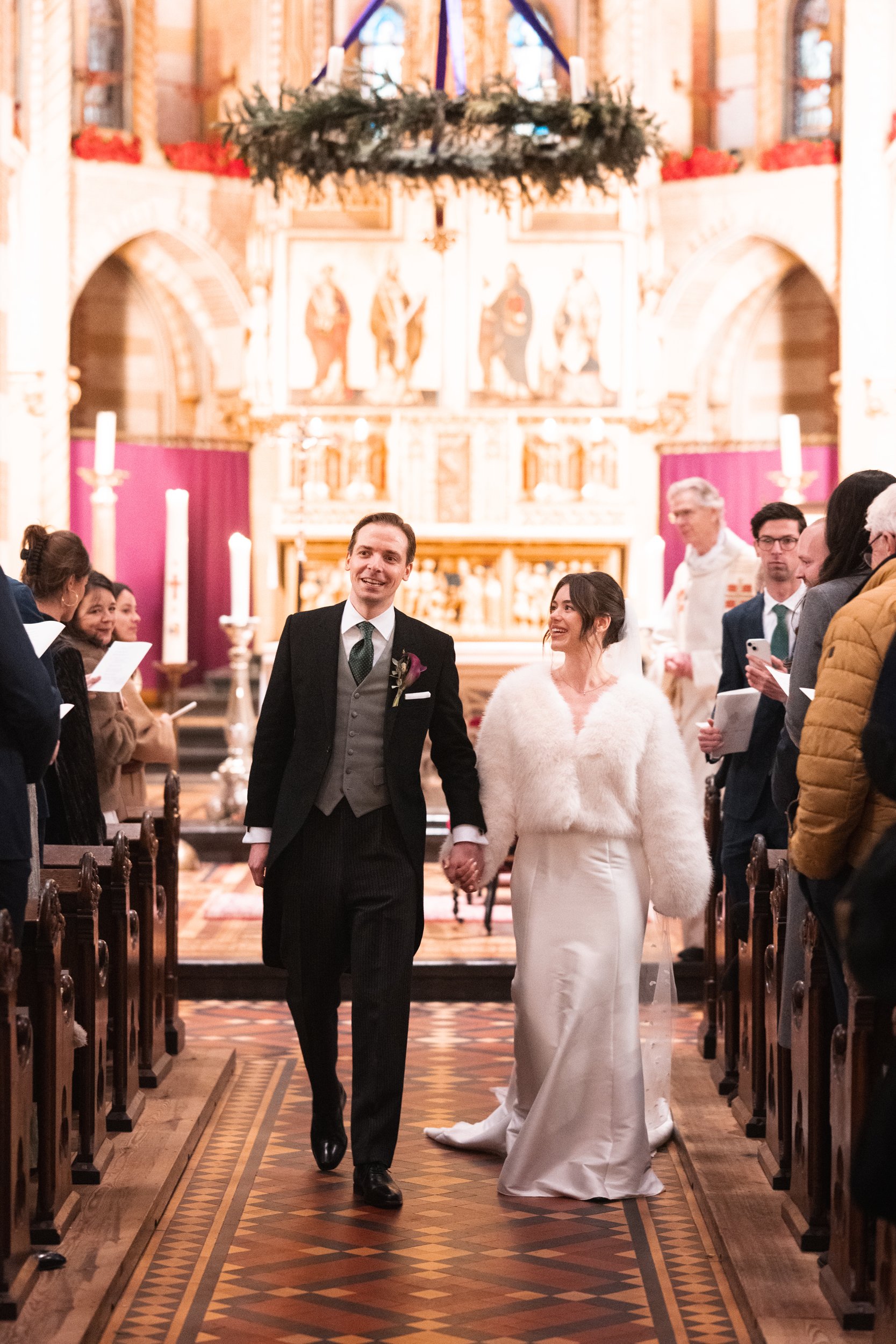 A newlywed couple walking down the aisle of a church after their wedding ceremony, surrounded by guests, clergy, and church decorations.