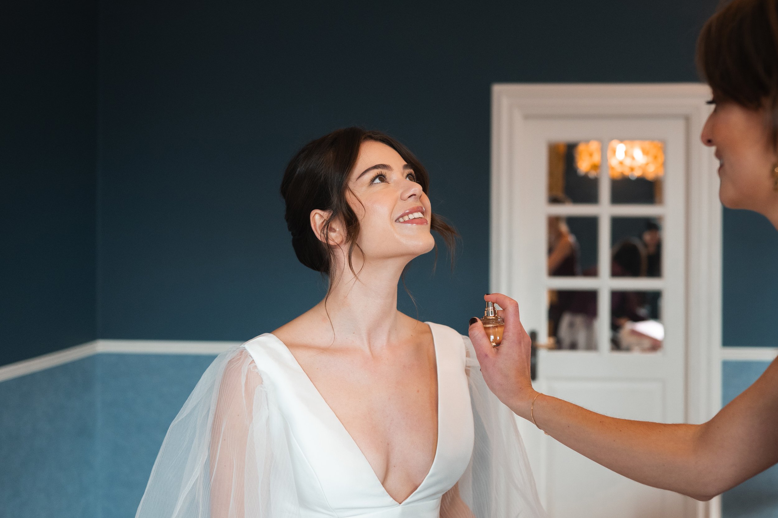 Bride smiling as her friend applies perfume in a room with blue walls and a decorative mirror.