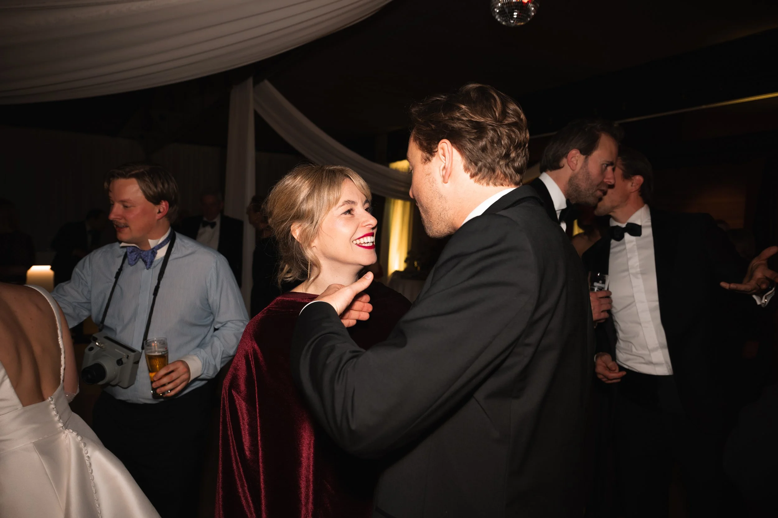 A woman and man dance closely at a formal event, smiling at each other. The woman is wearing a red dress and the man a black tuxedo. Other attendees in tuxedos are visible in the background, with some holding drinks.