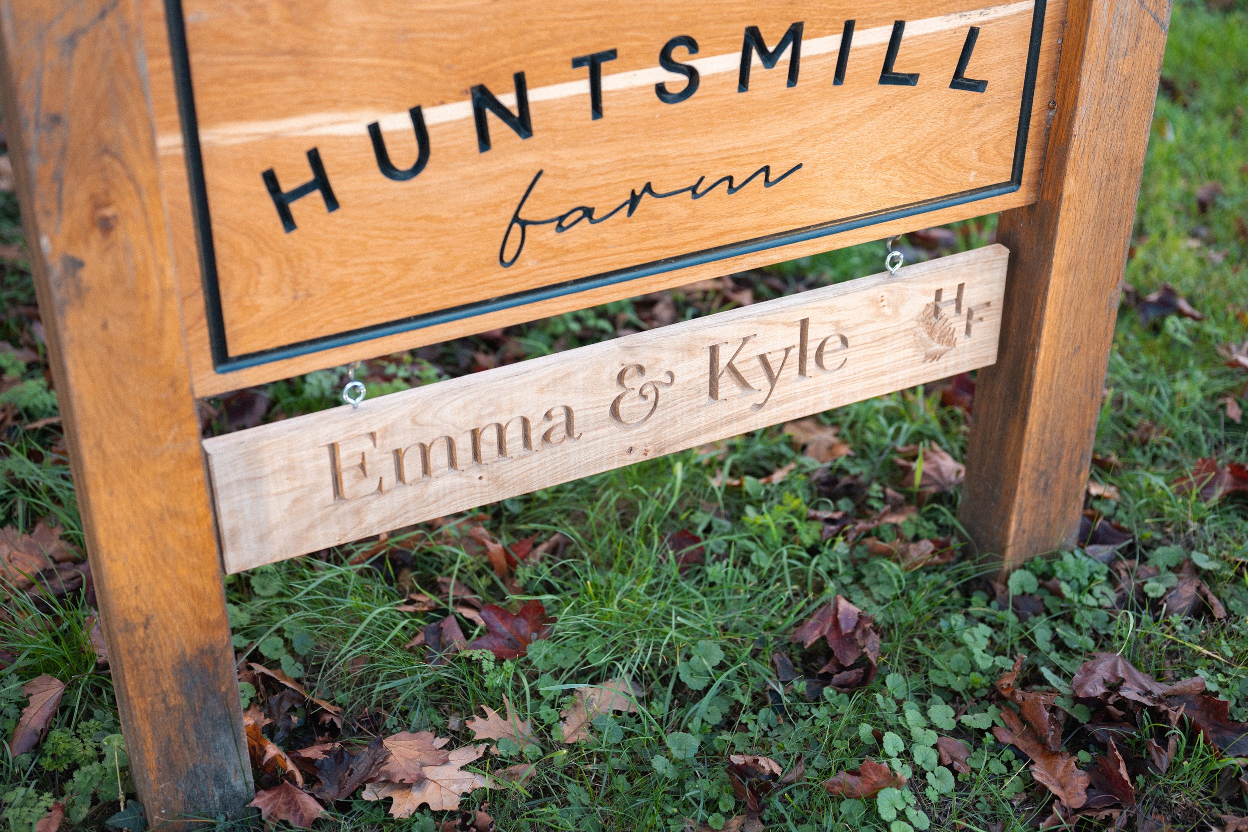 Wooden farm sign that reads 'HUNT SMILL farm' with a smaller wooden sign below engraved with 'Emma & Kyle' and a plus sign, surrounded by grass and fallen leaves.