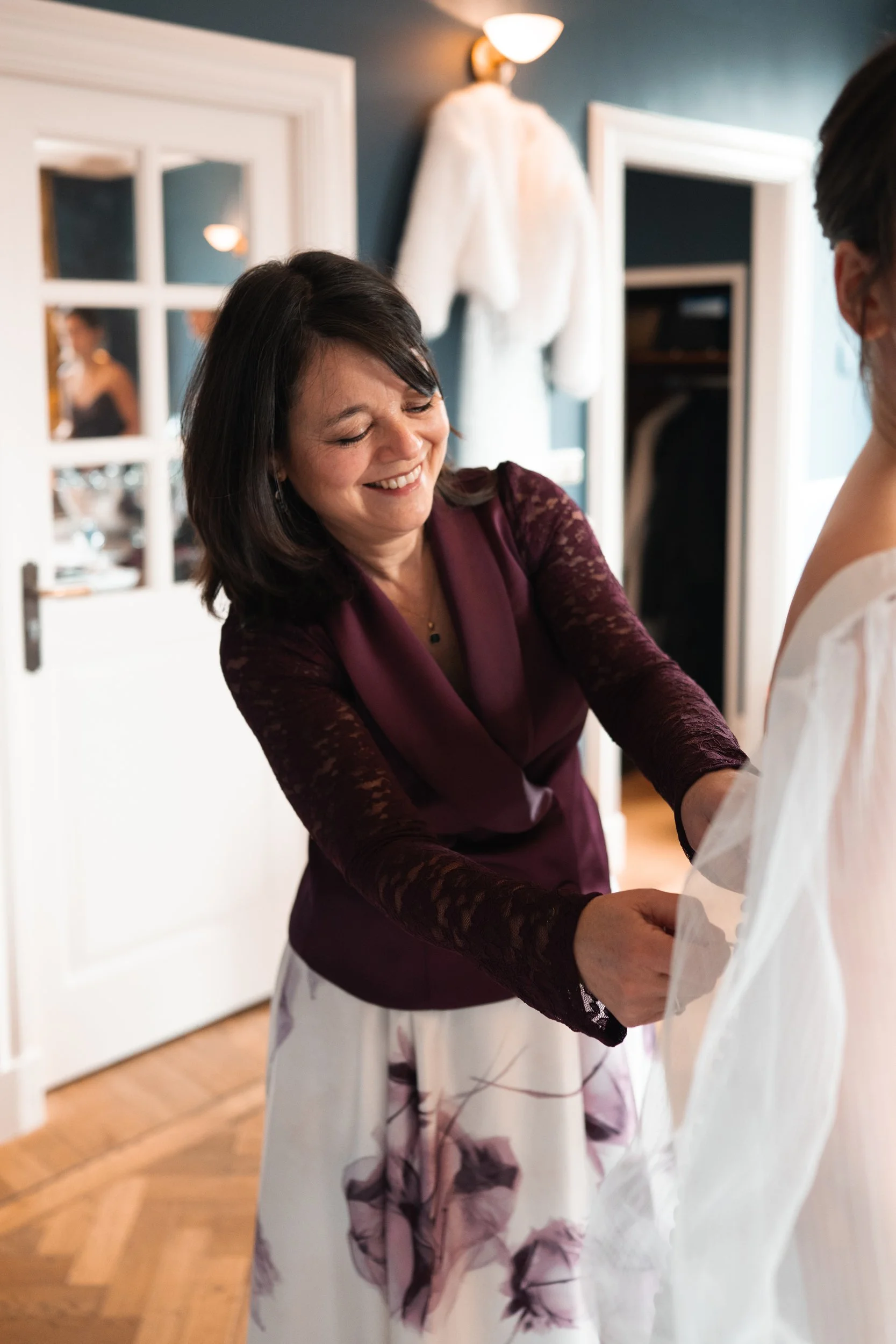 A woman smiling while helping a bride with her wedding dress indoors.