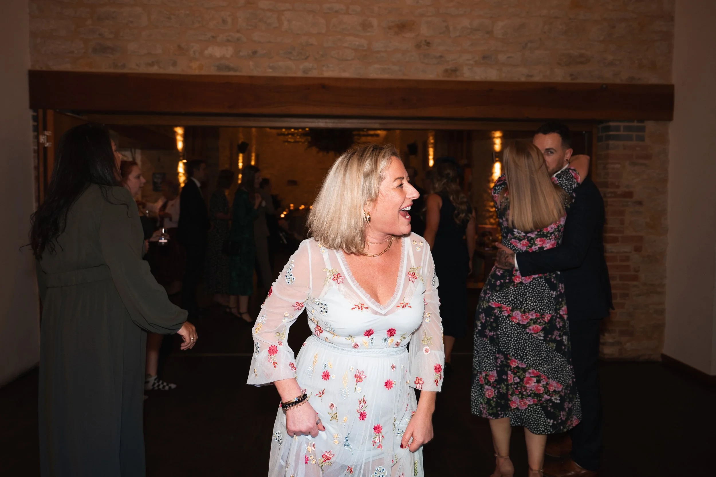 A group of people dancing and socializing at an indoor party or celebration. The scene features a middle-aged woman laughing and enjoying herself, wearing a white dress with embroidered flowers. In the background, couples are dancing, including a man