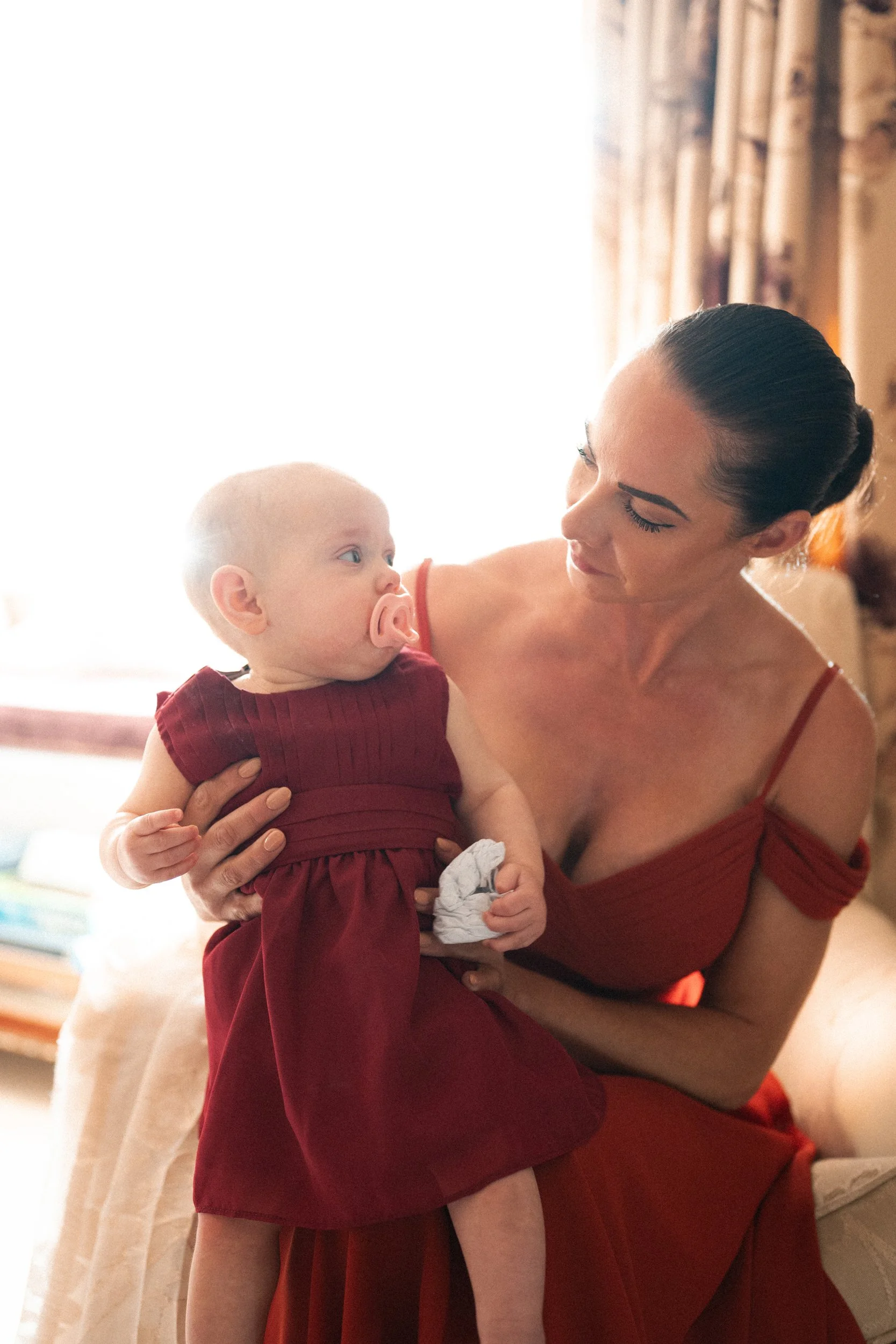 A woman in a red dress holding a baby with a pacifier, sitting on a beige couch in a well-lit room.