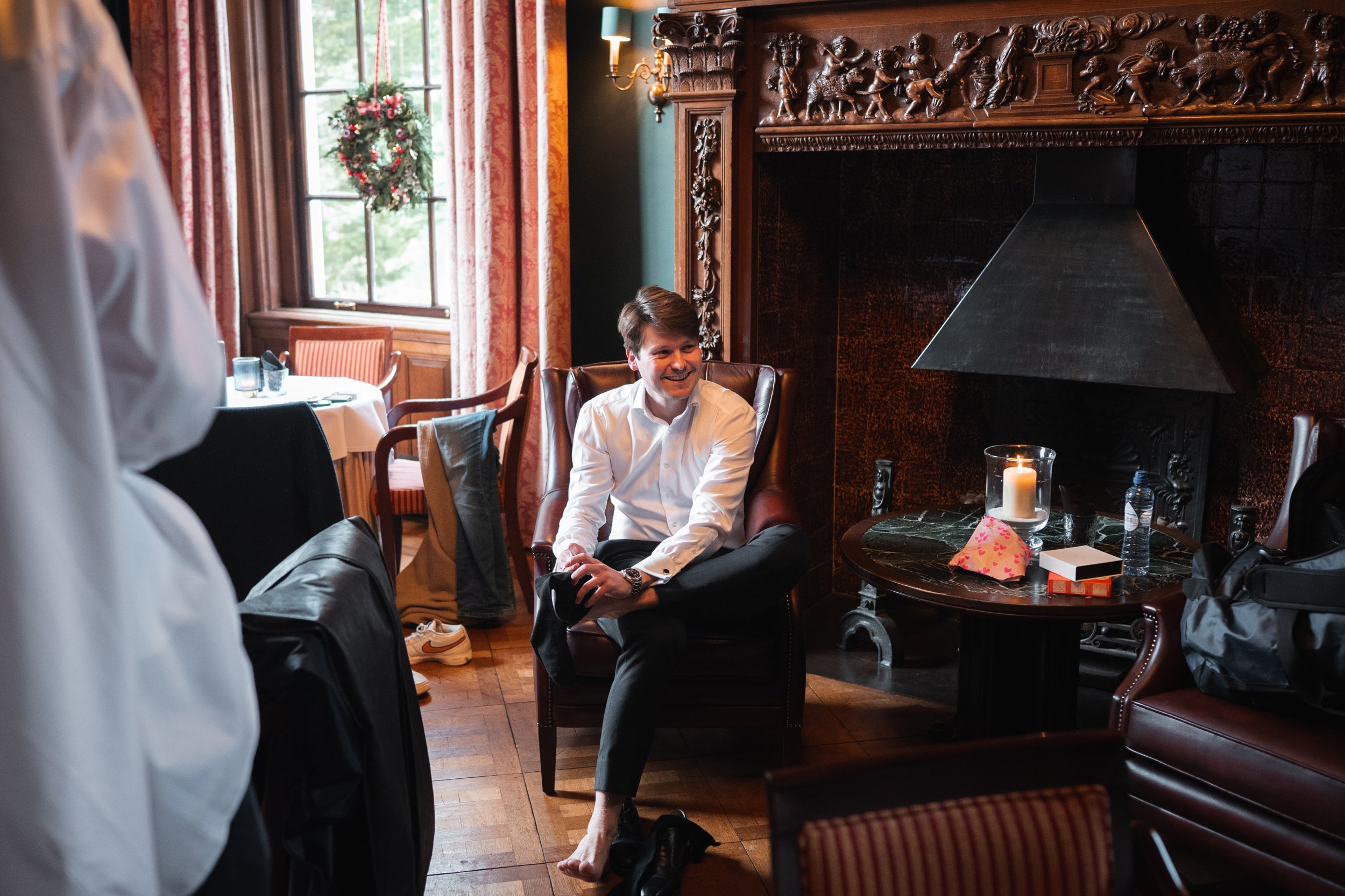 A smiling man in a white shirt sitting barefoot in a leather armchair in a warmly decorated room, with a fireplace, lit candle, and holiday wreath visible.