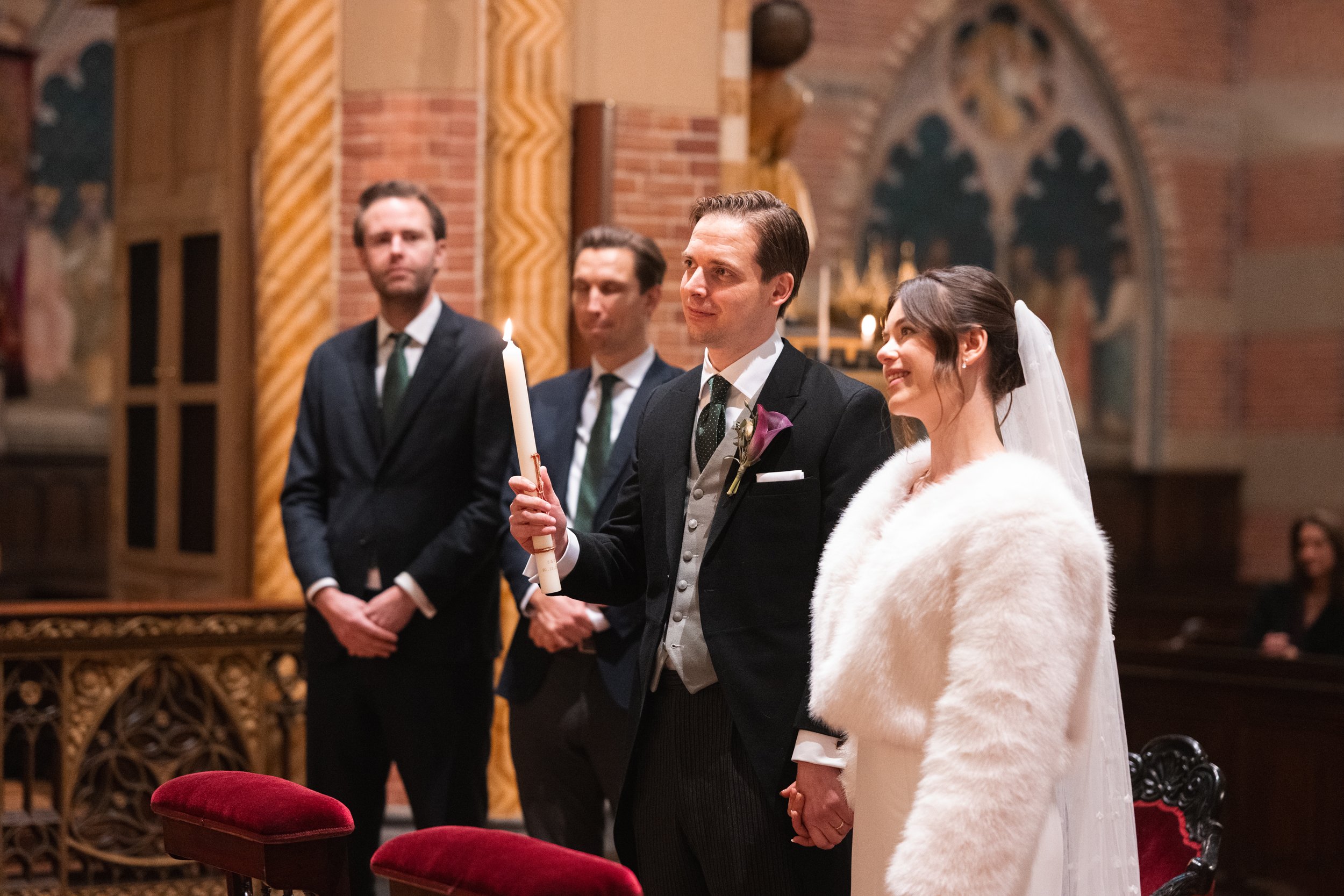 A wedding ceremony inside a church with the bride and groom holding hands, the groom holding a lit candle, and two men standing behind them.