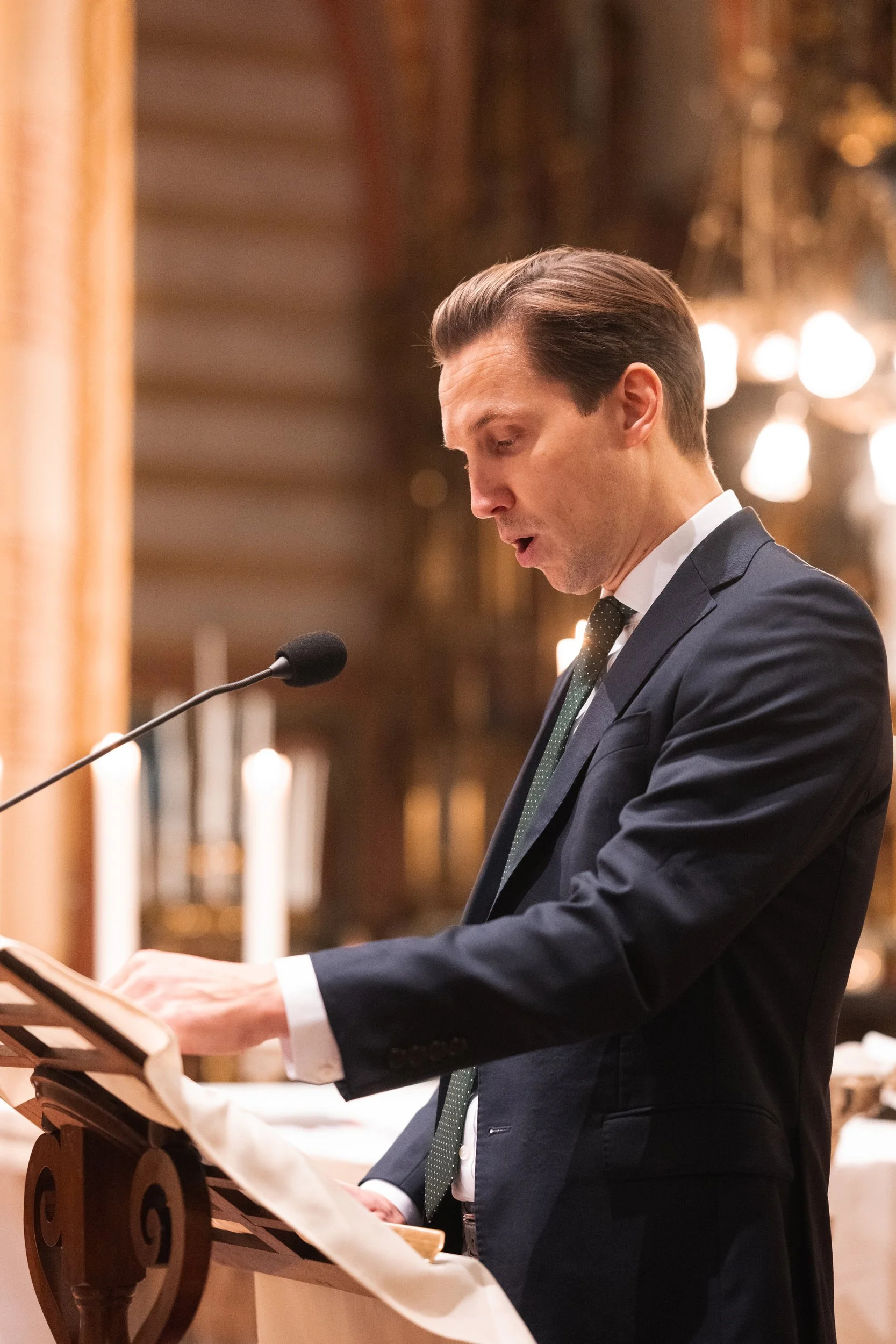 A man in a dark suit and tie stands at a lectern, reading or speaking into a microphone in an ornate, warmly lit room with candles and decorative chandeliers.