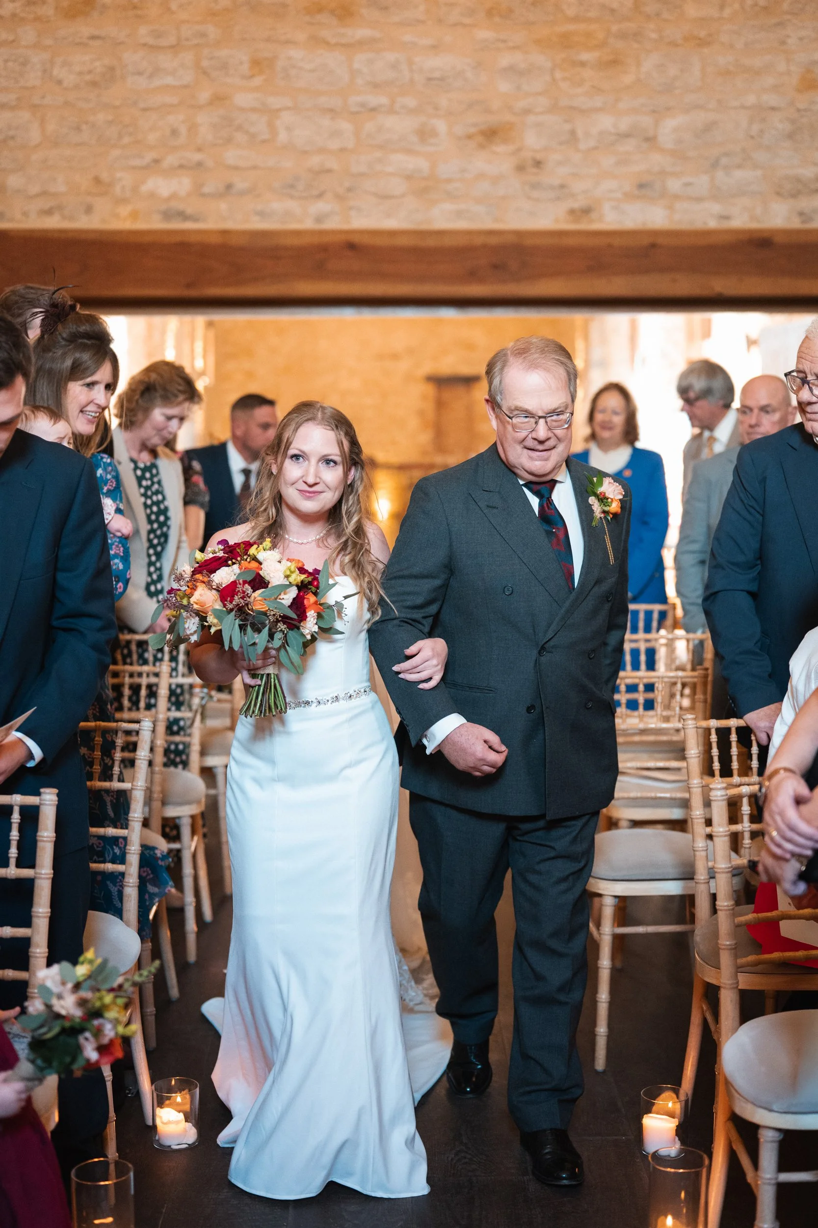 A bride walking down the aisle with an older man, likely her father, at a wedding ceremony. The bride is holding a bouquet and wearing a white wedding dress, while the man is dressed in a dark suit. Guests are seated and standing on either side, watc