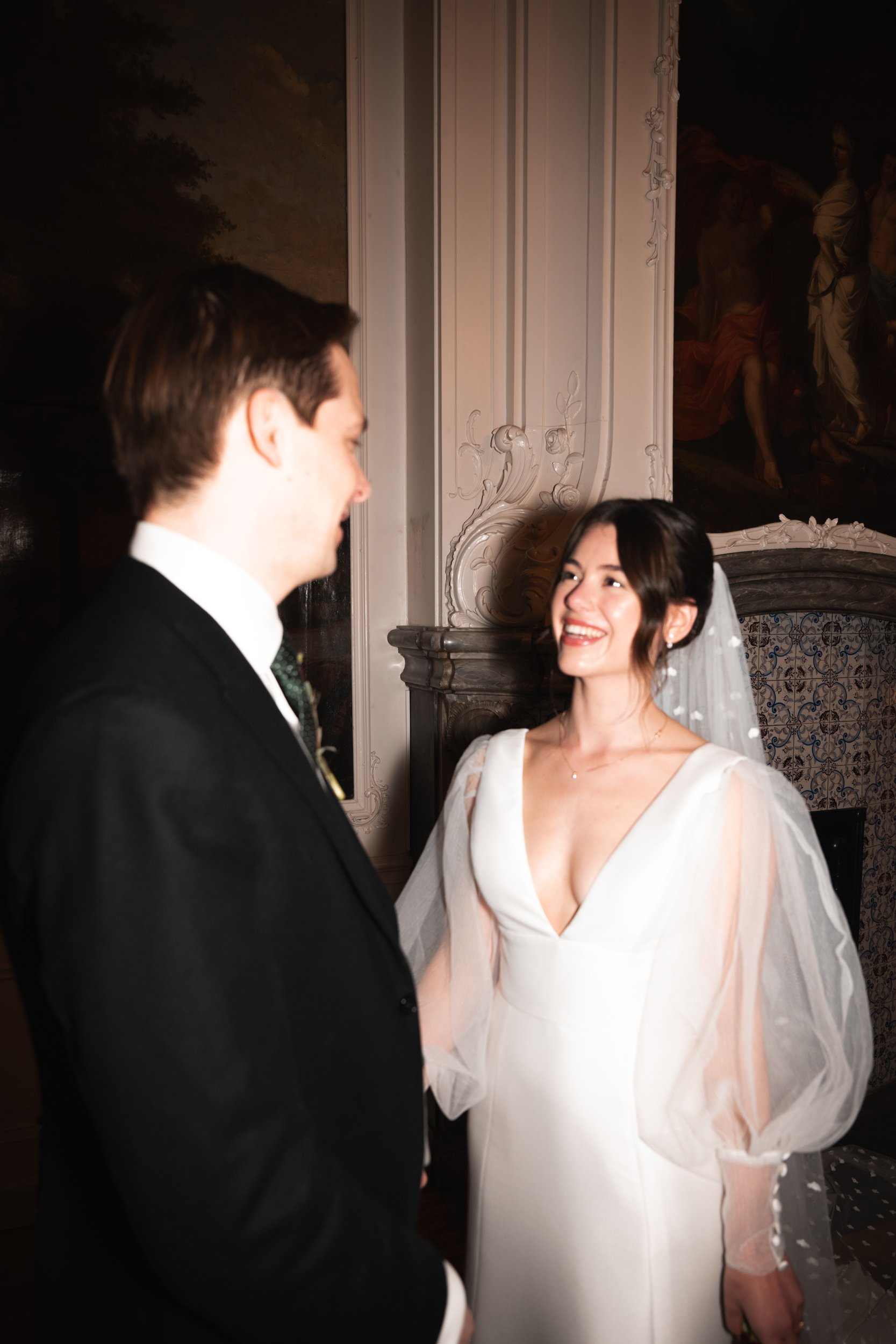 Bride and groom smiling at each other during wedding ceremony indoors with ornate walls and classical paintings in the background.