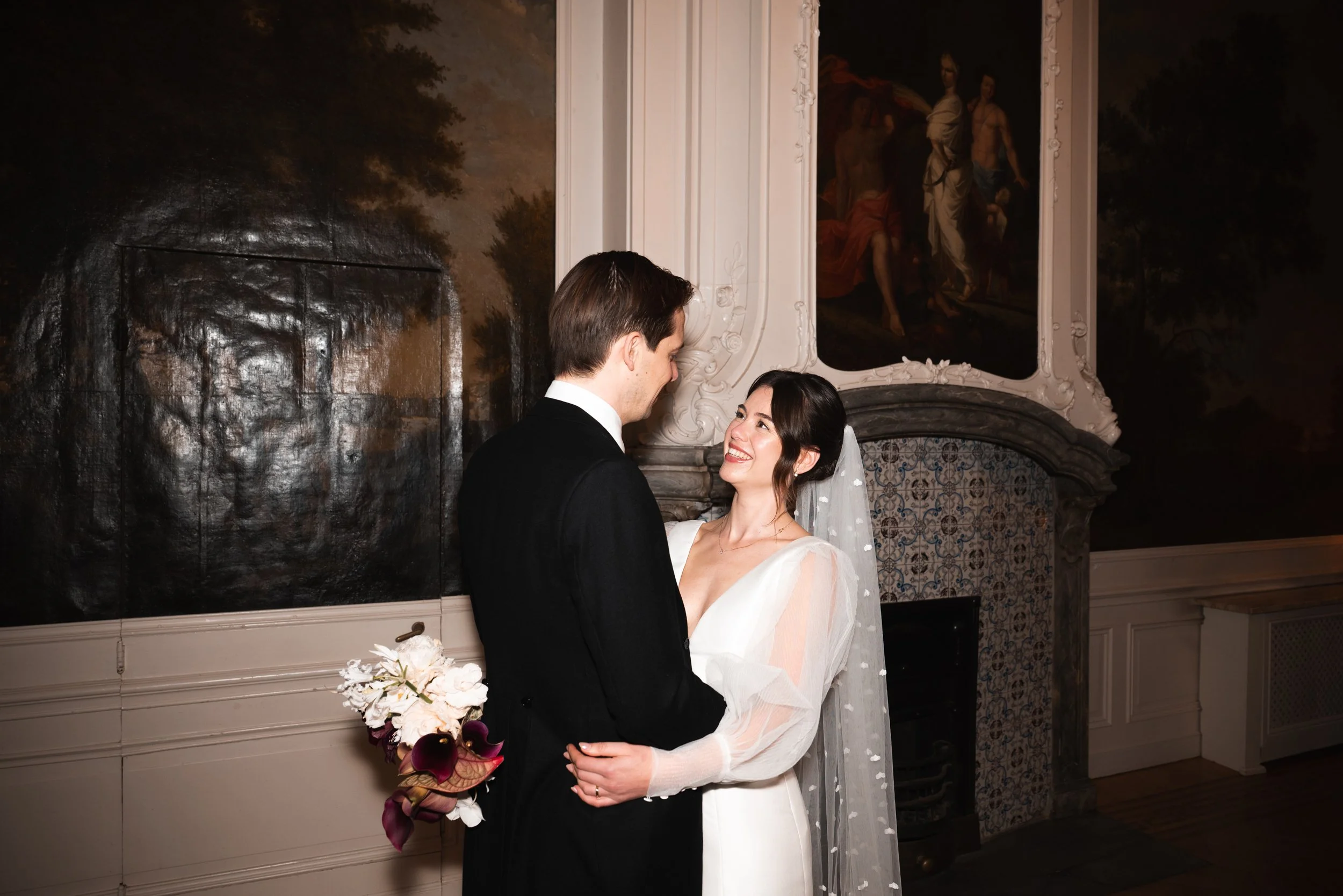 A bride and groom smiling at each other in an indoor setting with ornate decor, a fireplace, and large classical paintings on the wall.