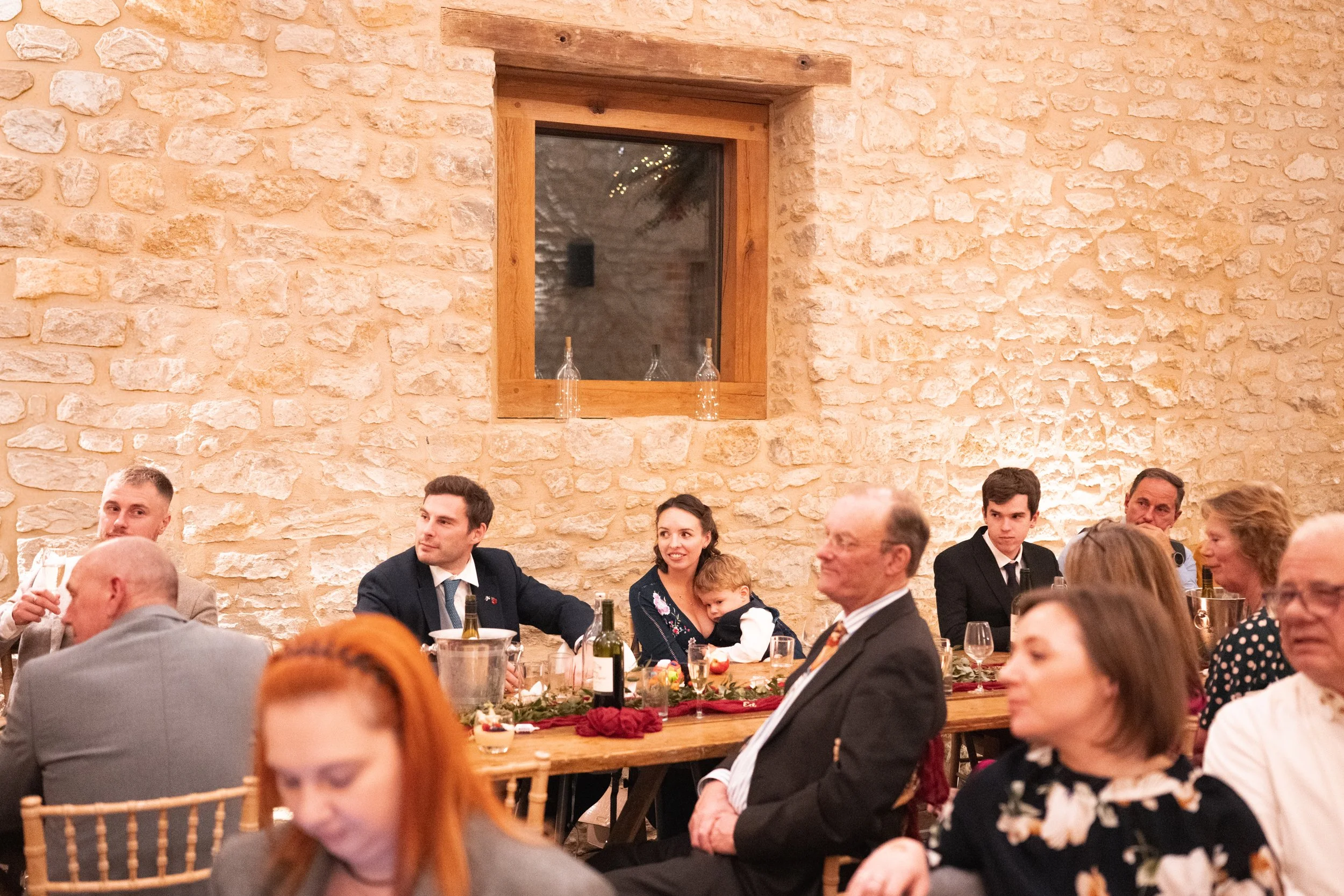 People sitting at a long table during a social gathering in a rustic indoor setting with a stone wall and a window.