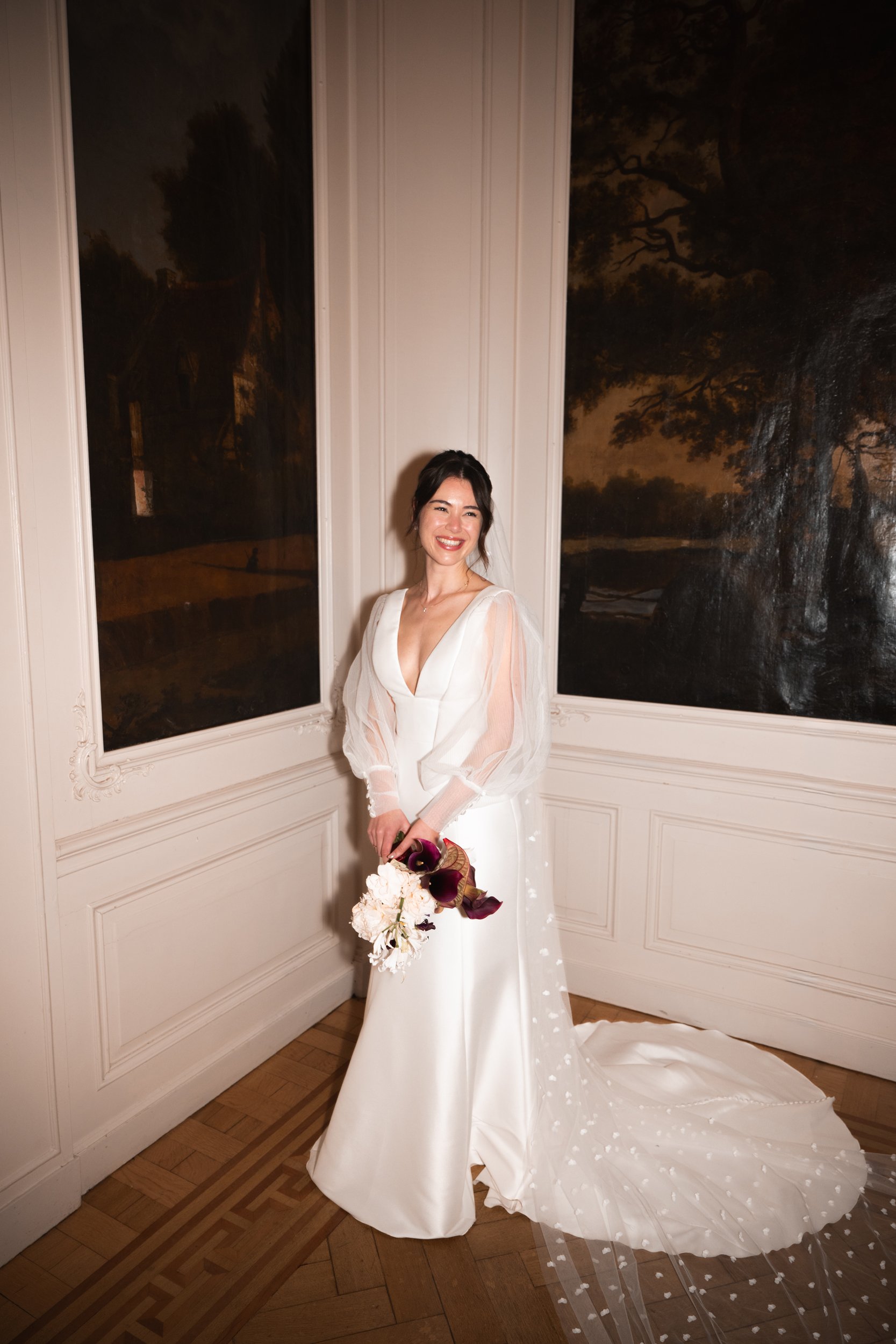A woman in a white wedding dress holding a bouquet of dark purple and white flowers, standing indoors near large windows with dark landscape paintings.