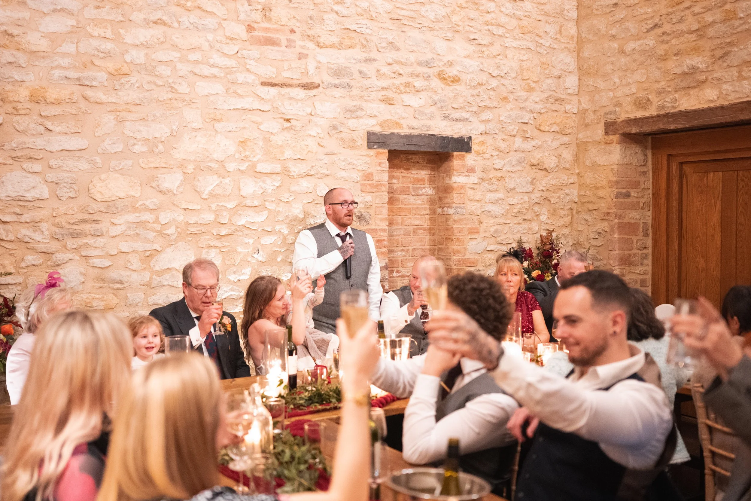 People gathered at a wedding reception, with a man giving a speech at a decorated table against a brick wall.