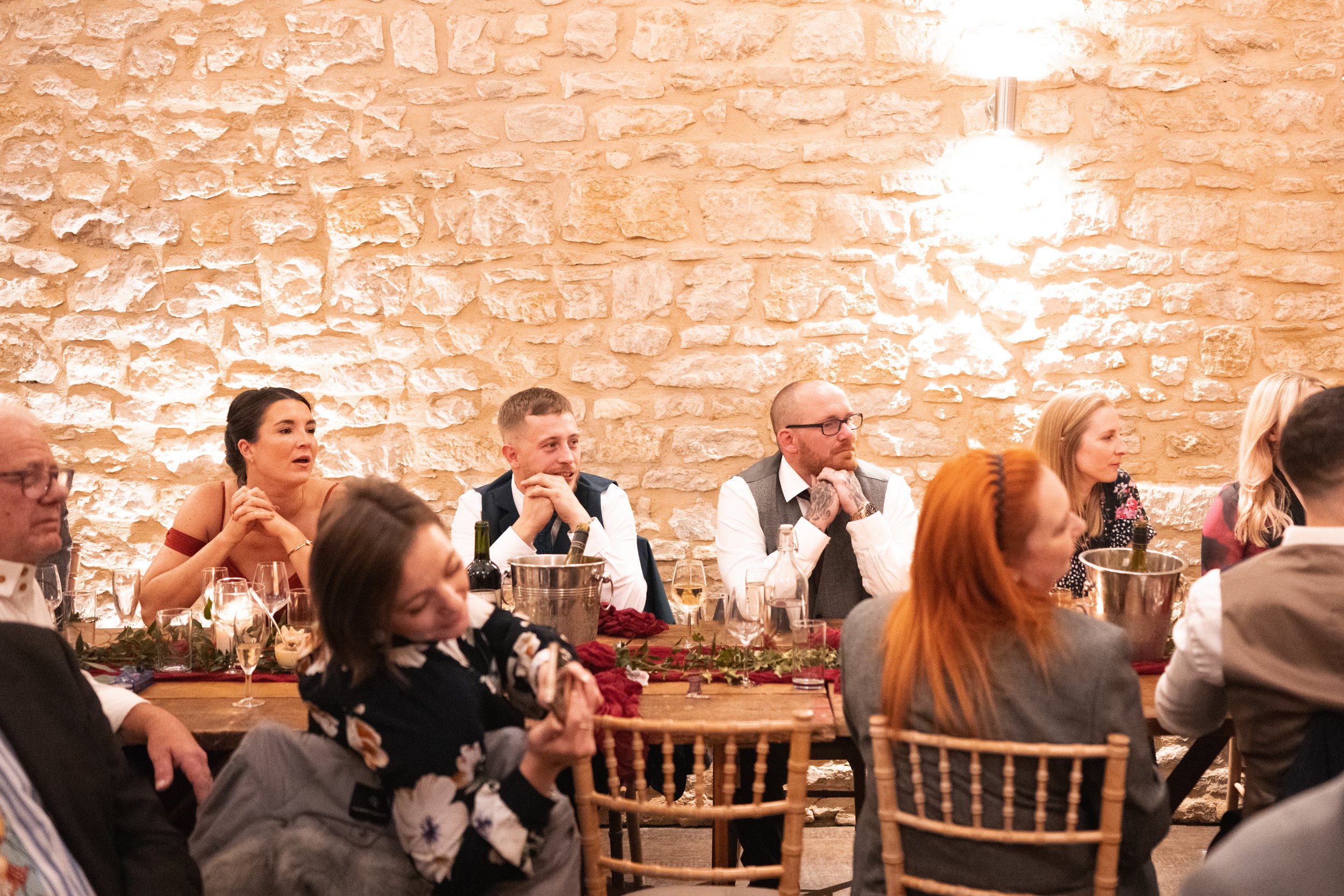 People sitting at a long wooden table during a formal event or dinner in a room with a beige brick wall.