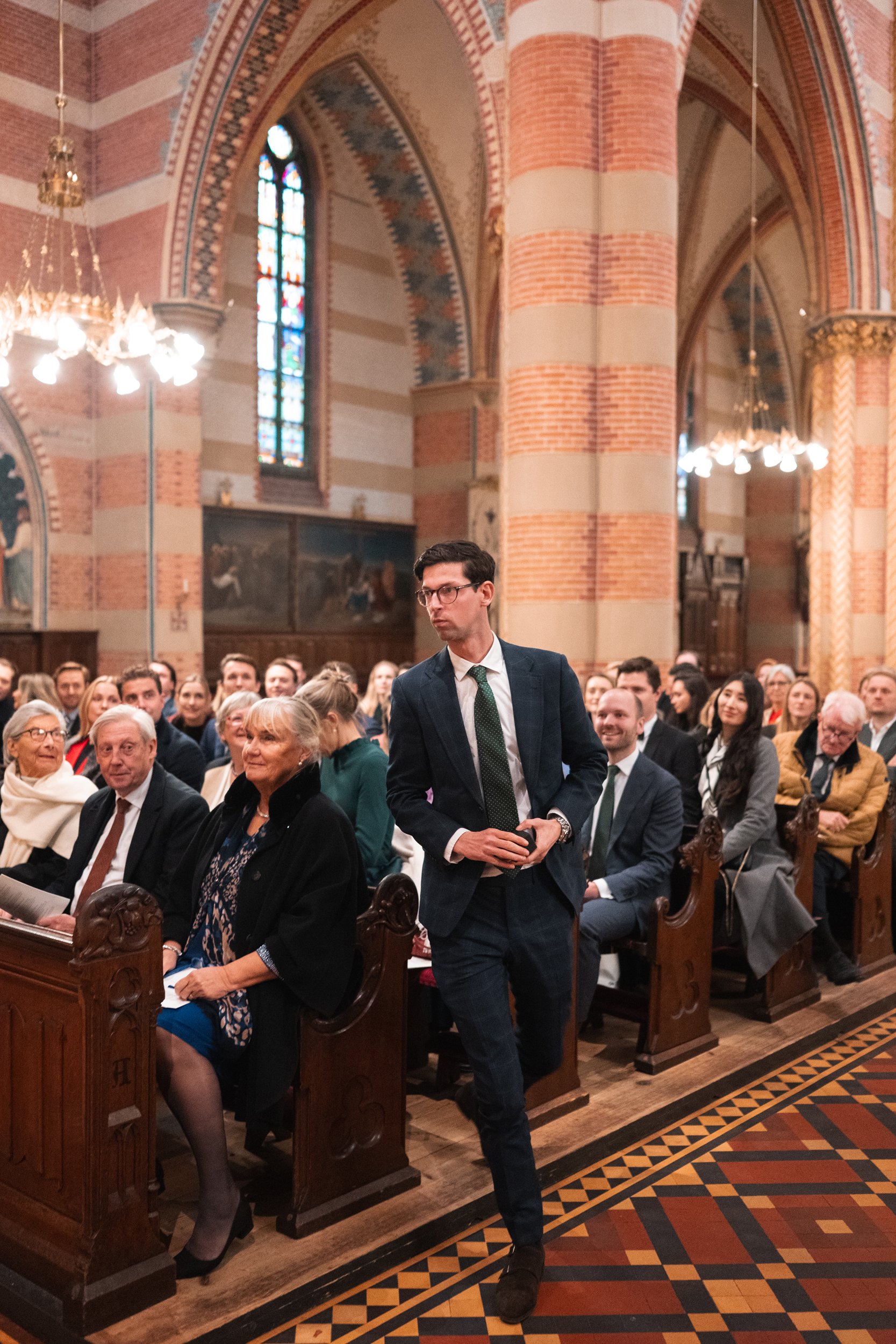 A man in a dark suit and glasses walking past a seated congregation inside a church with stained glass windows and brick walls.