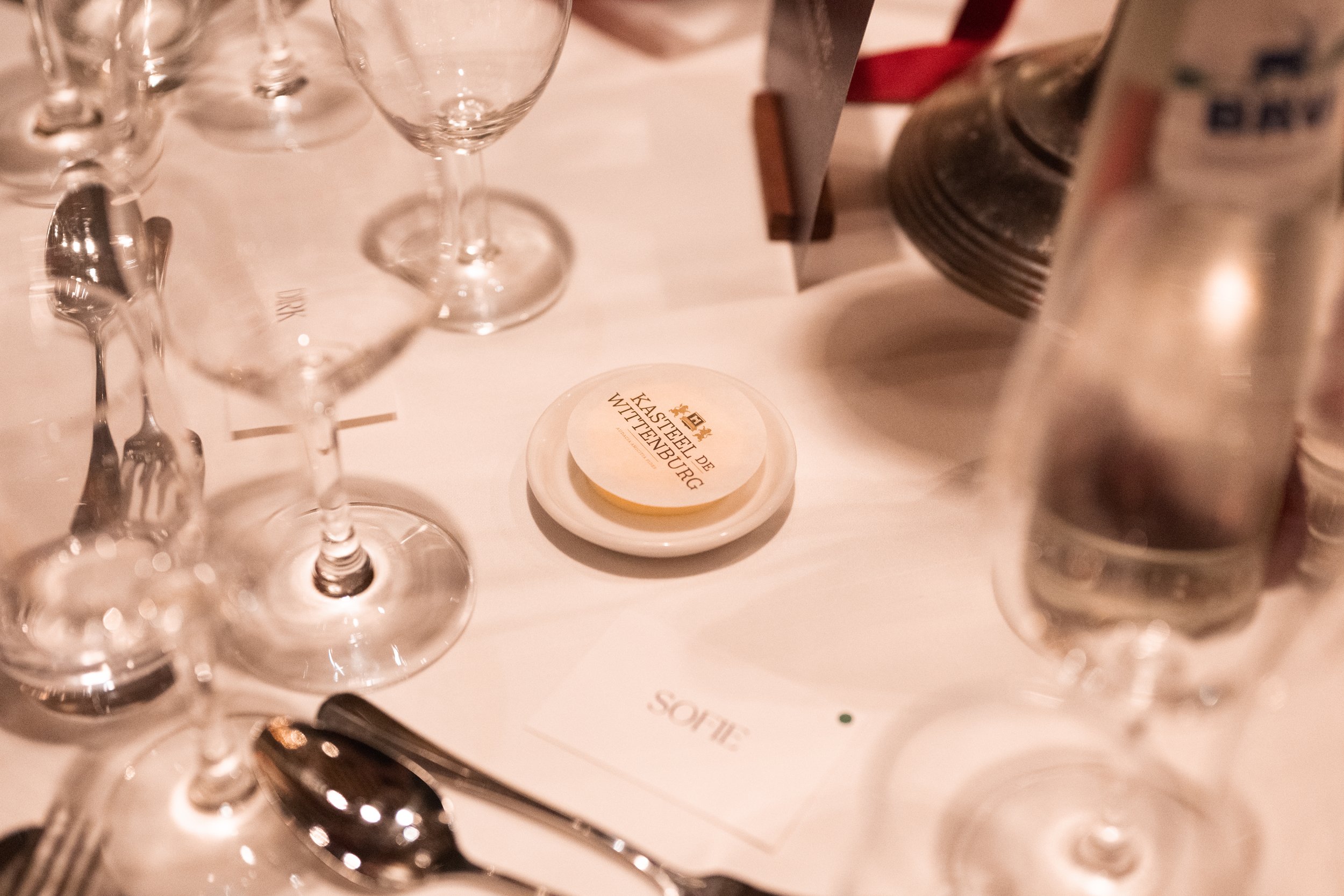 Empty wine glasses, a butter dish with a paper wrapper labeled 'KAASSTEL DE WITTE ENBURG,' a water glass containing water, and silverware on a table with a white tablecloth, possibly in a restaurant setting.