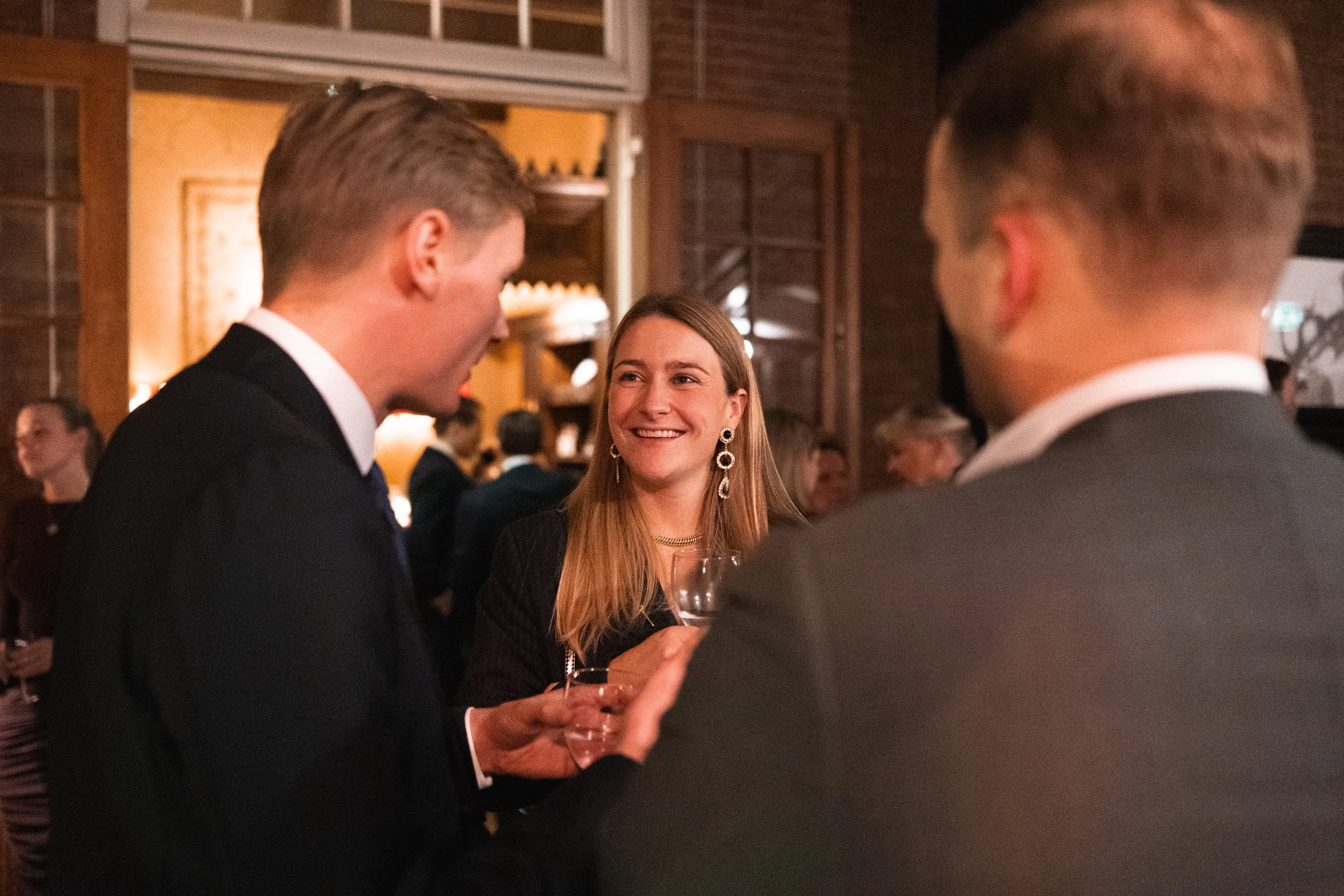 A woman smiling, holding a wine glass, engaged in conversation with two men at a social event or party.