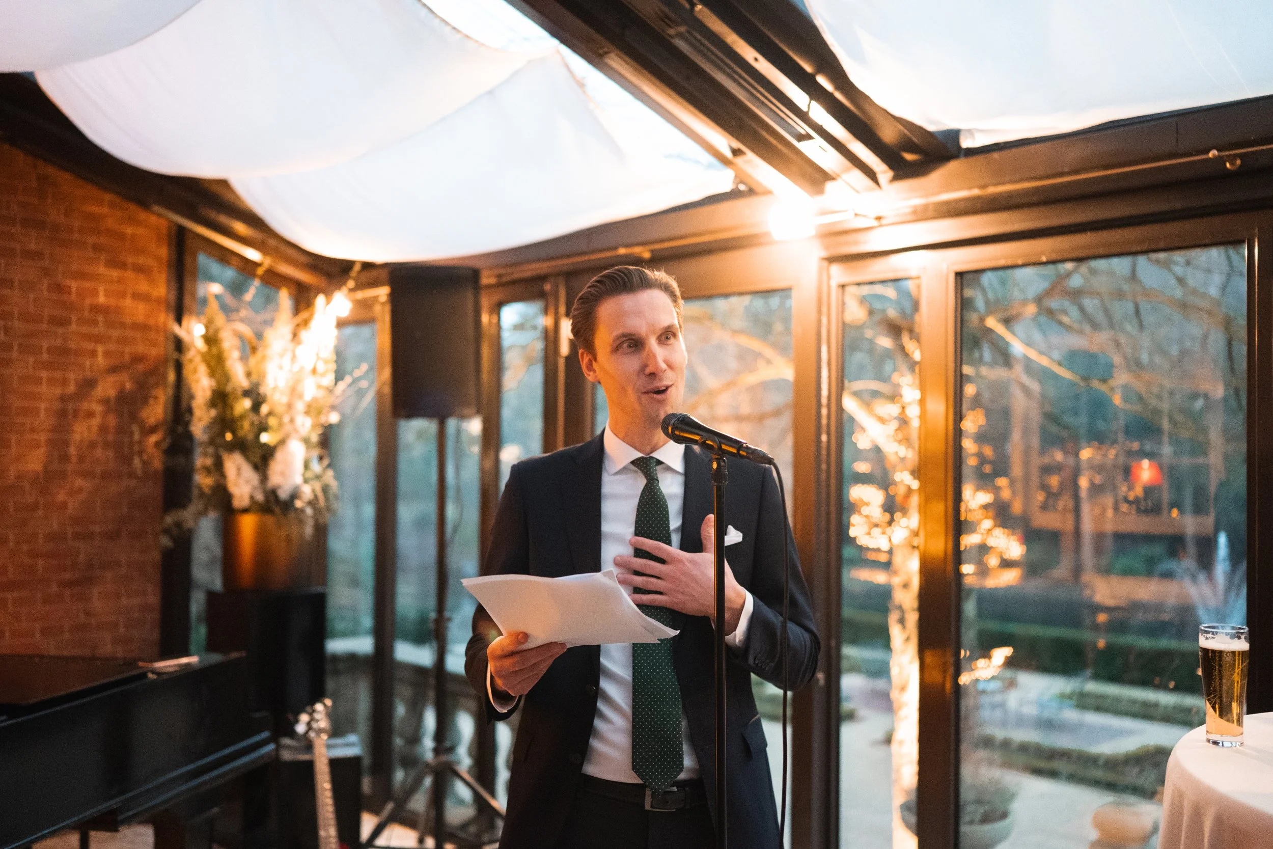 A man in a dark suit and tie giving a speech at a microphone with papers in hand, in a warmly lit indoor space with large windows and outdoor scenery.
