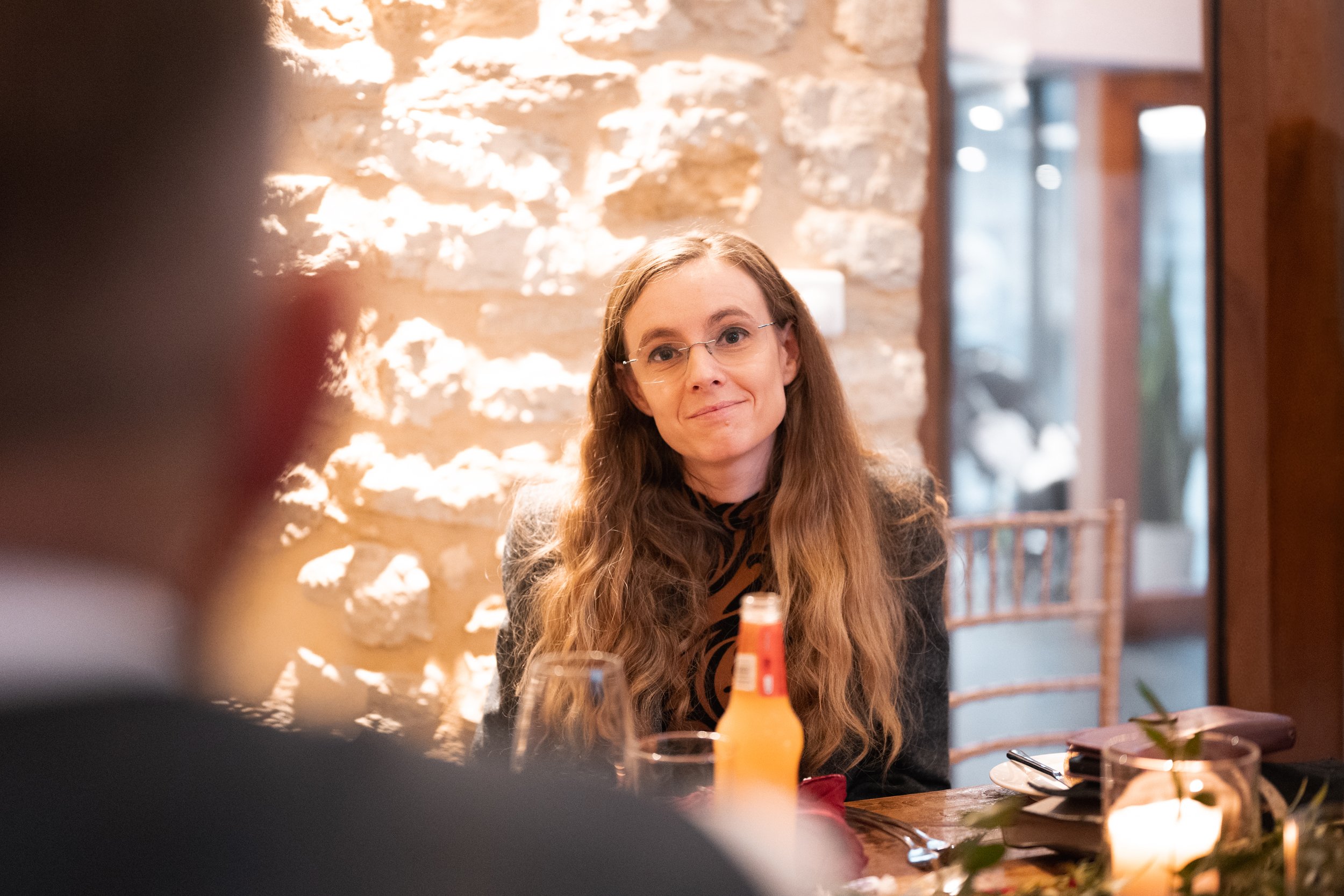 A woman with long wavy hair, glasses, and a grey blazer sitting at a restaurant table. She has a slight smile and is looking towards the camera. The table has a bottle of orange-colored drink, a plate, cutlery, and a small candle. The background has 
