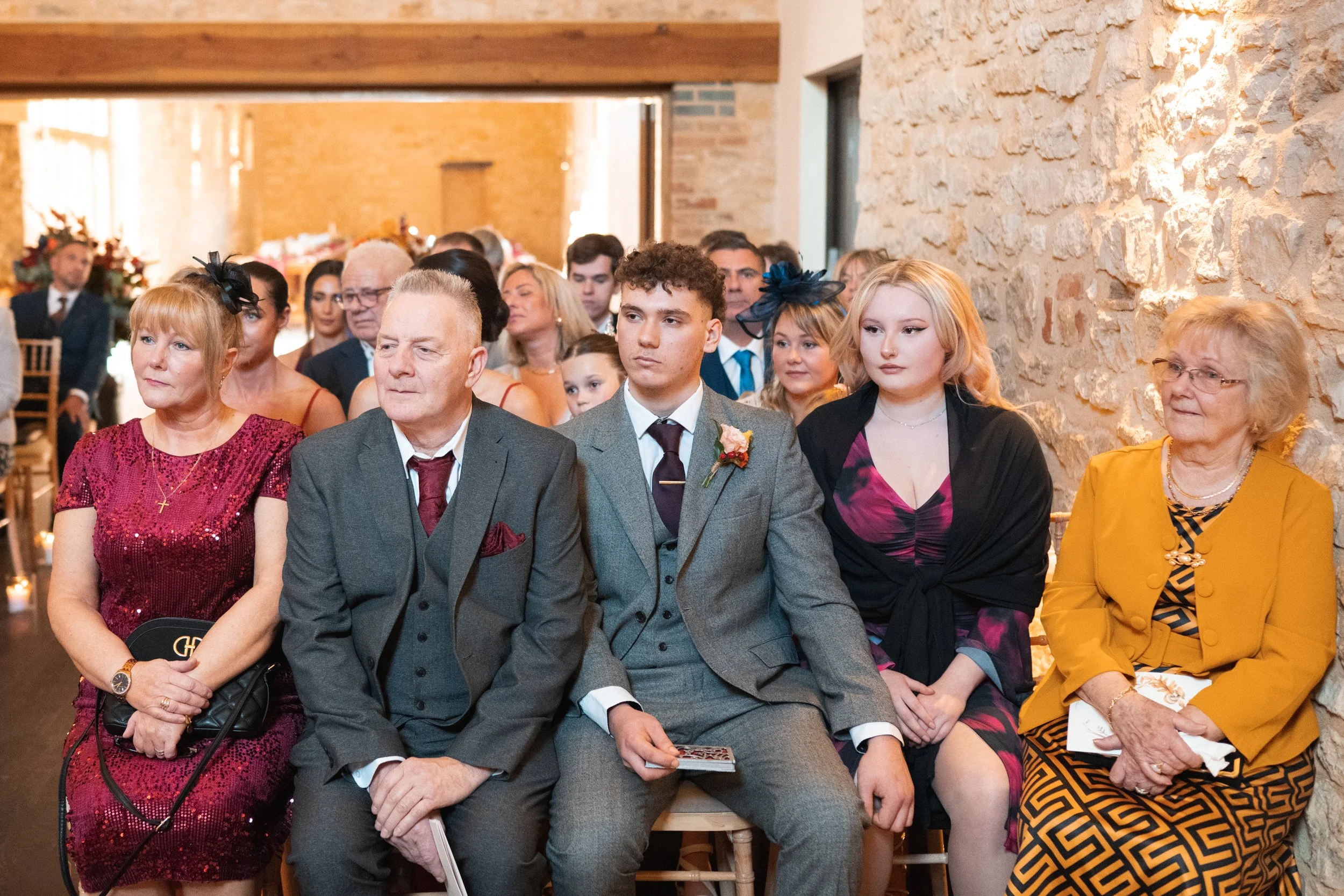 Group of people attending a formal event seated on wooden chairs in a rustic venue with brick walls.