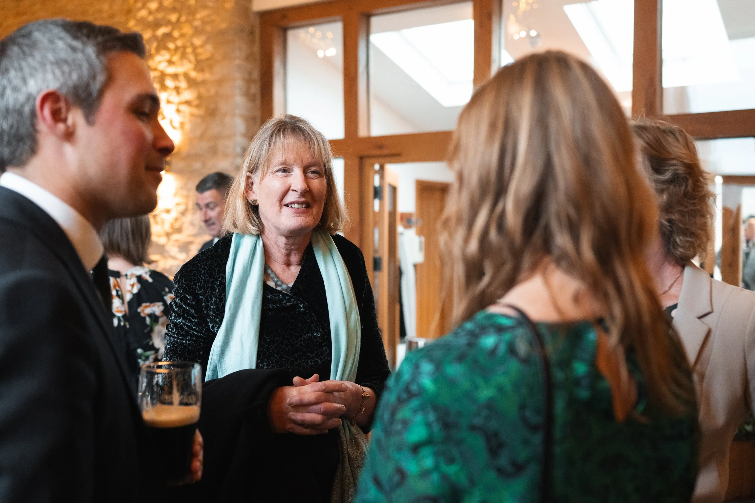 A group of people at a social gathering indoors, engaged in conversation, with a woman in the center smiling and talking to others.