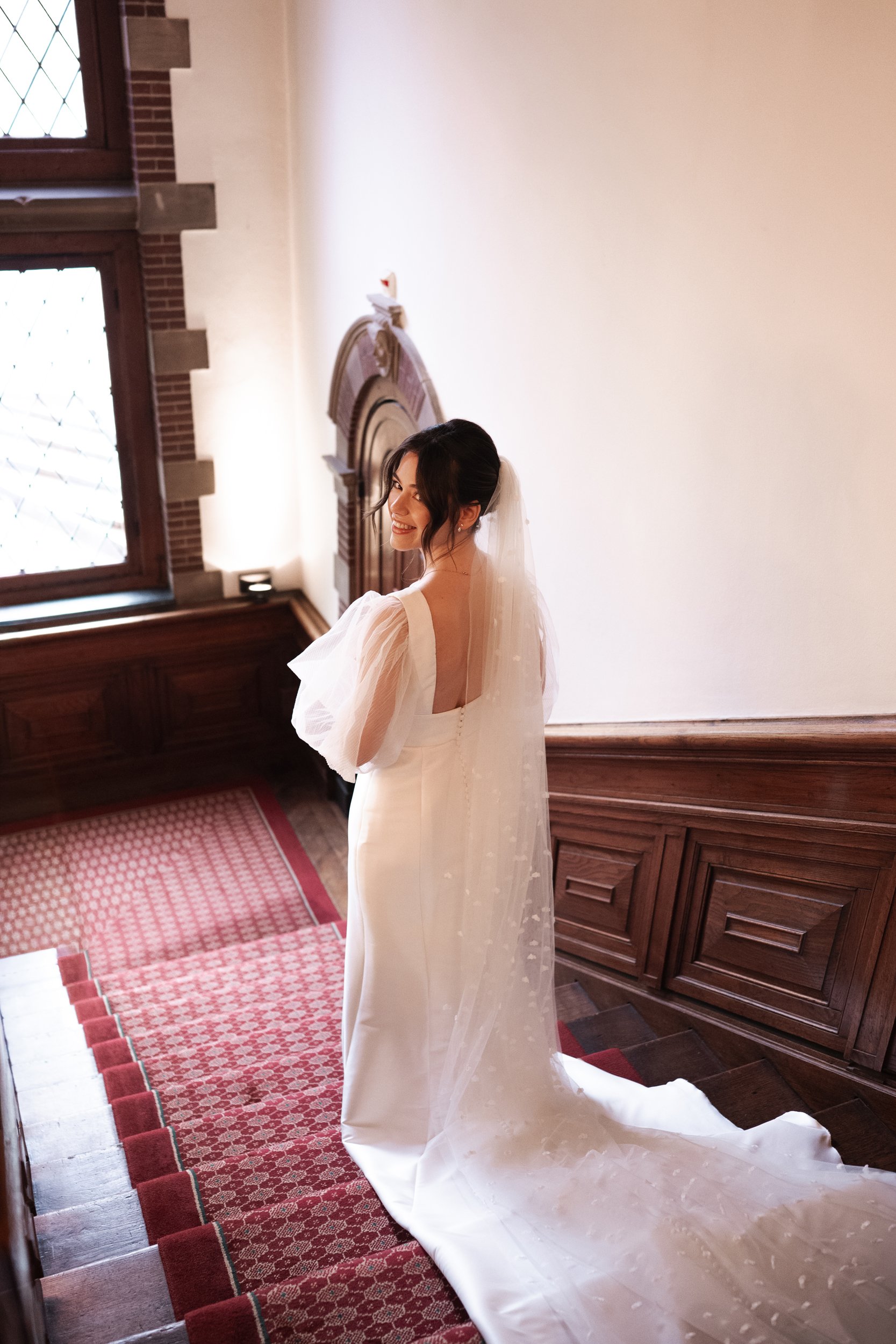 A bride in a white wedding dress with a long train and veil, standing on a staircase inside a historic building with wooden paneling and a stained glass window, smiling and looking over her shoulder.