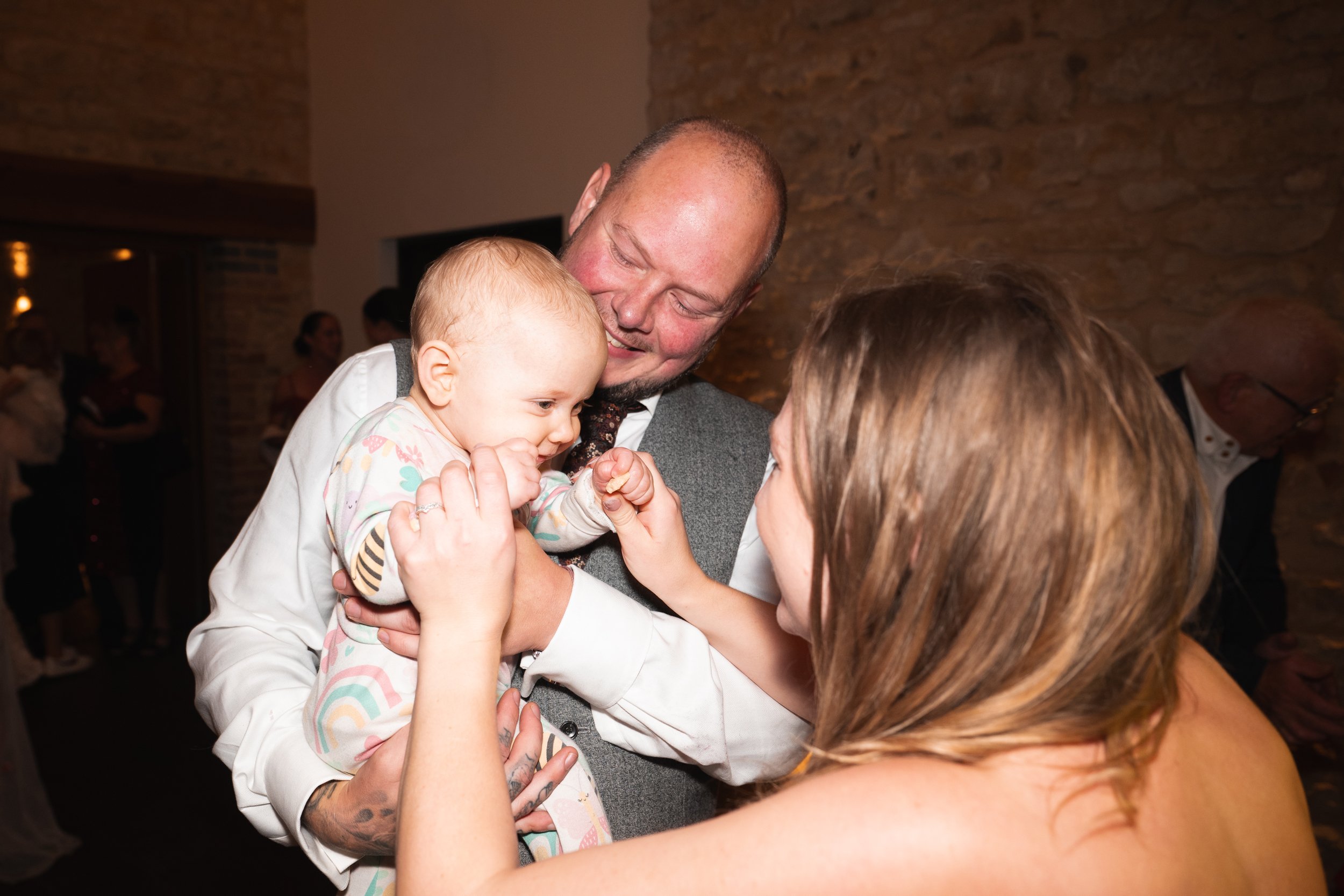 A man holding a baby, both smiling, as a woman playfully interacts with the baby in a warmly lit room with brick walls, in what appears to be a social gathering or celebration.