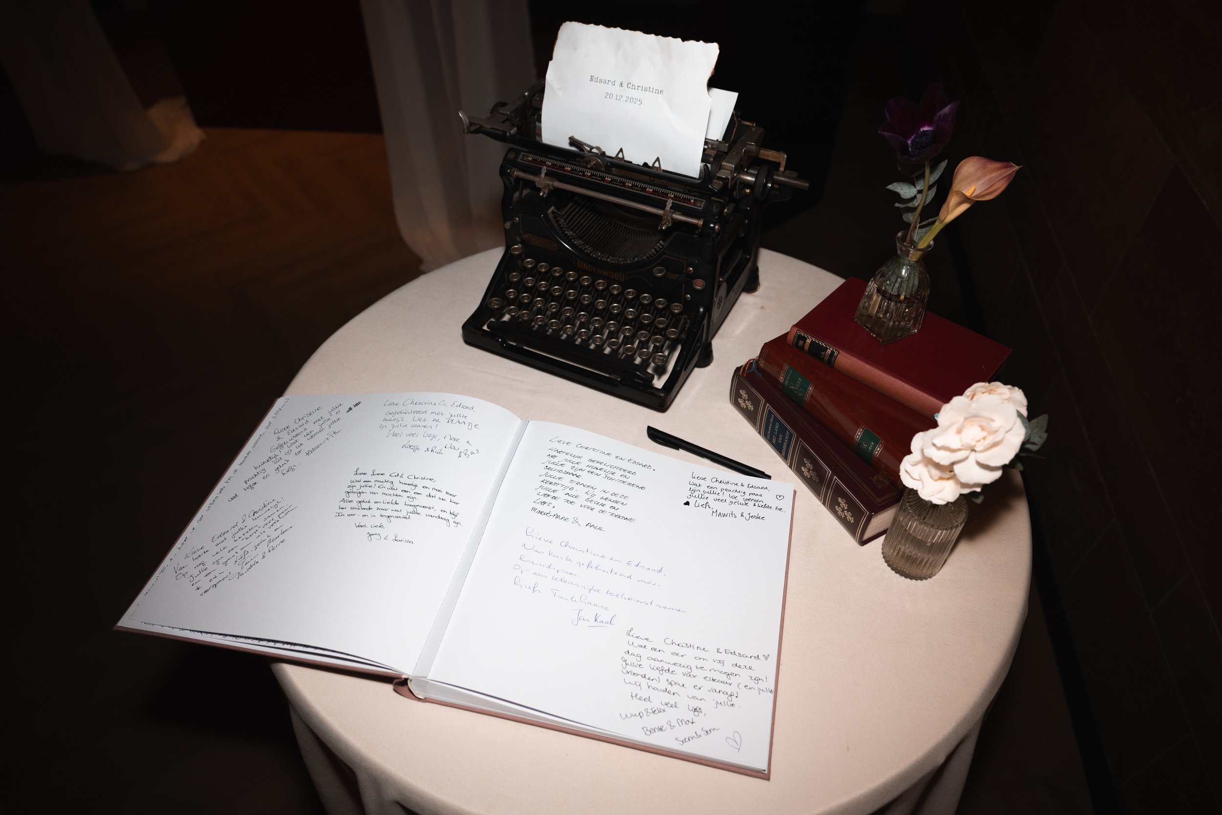 A vintage typewriter with a piece of paper, an open guestbook filled with handwritten messages, a stack of books, a pen, and two vases with flowers on a round table.
