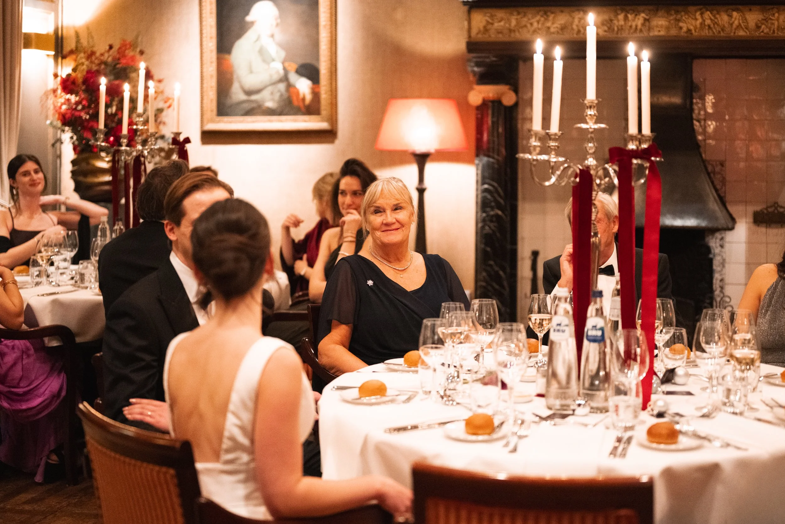 People dressed in formal attire seated around a dining table at a fancy dinner event, with a candelabra centerpiece, a fireplace, and artwork on the walls.