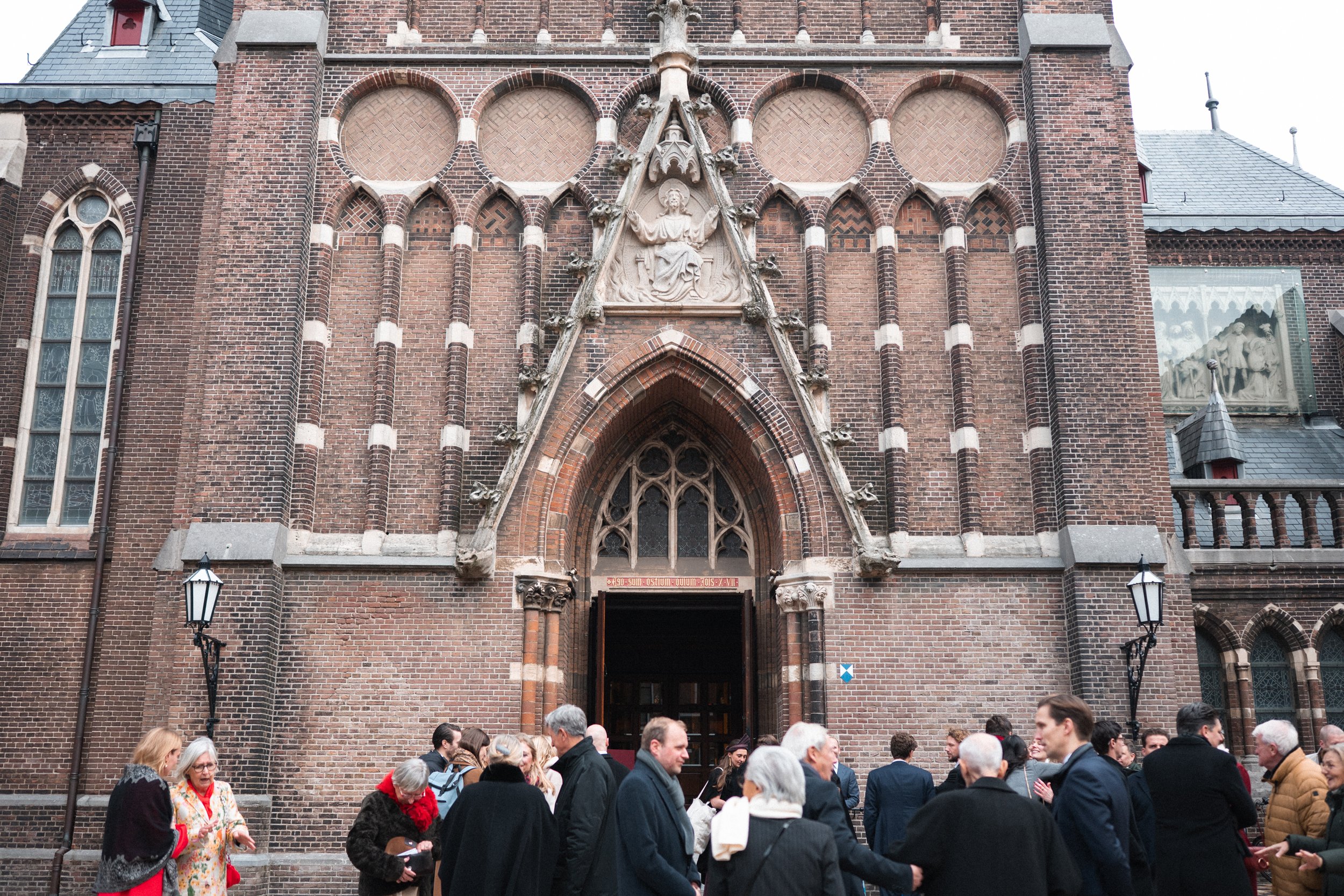 A group of people gathered outside a large brick church with gothic architectural features, including pointed arch doorway, stained glass windows, and intricate stone carvings above the entrance.
