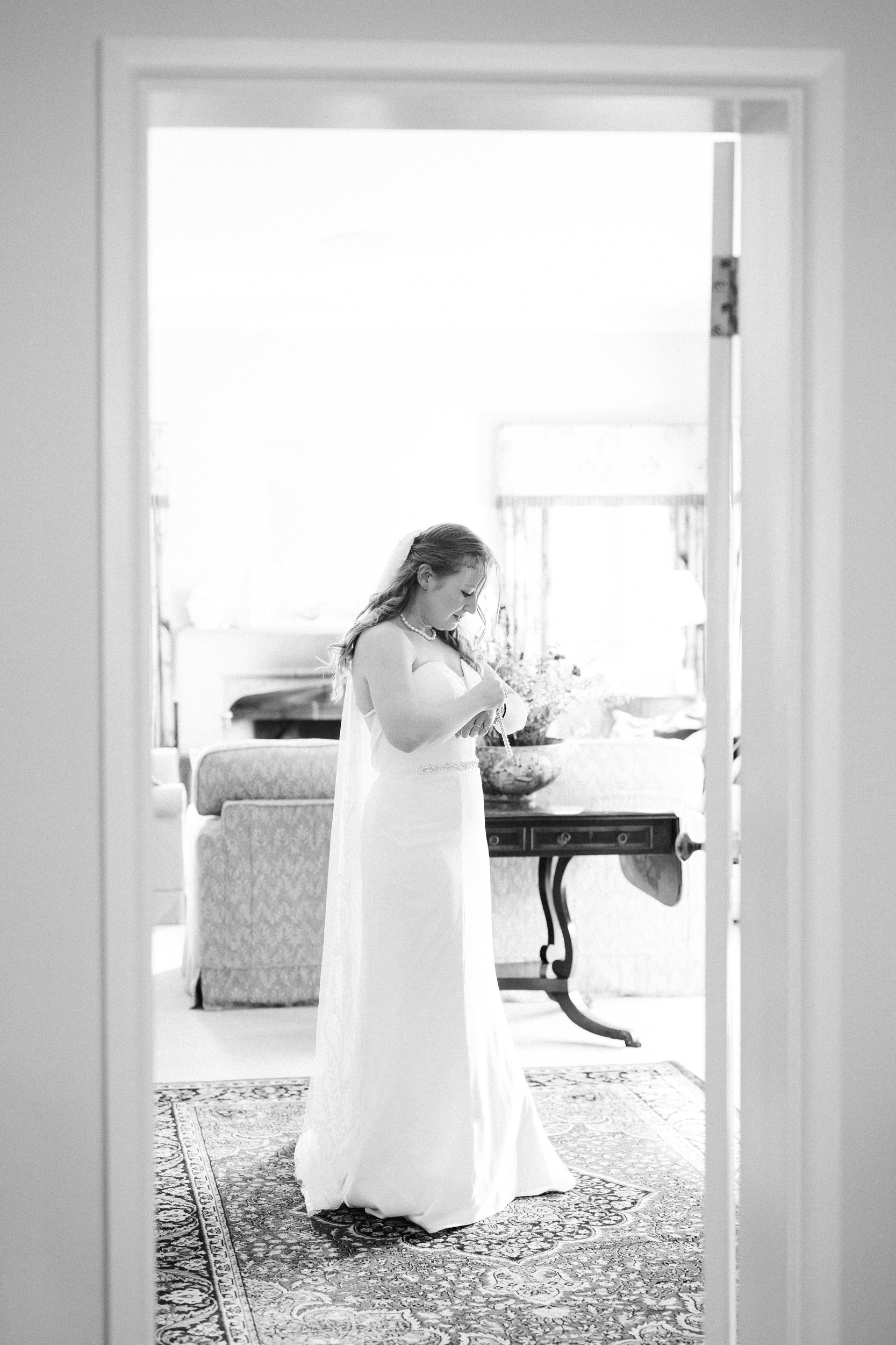 A bride in a white wedding dress standing indoors, looking down at her purse, with a floral arrangement in the background and a patterned rug under her feet.