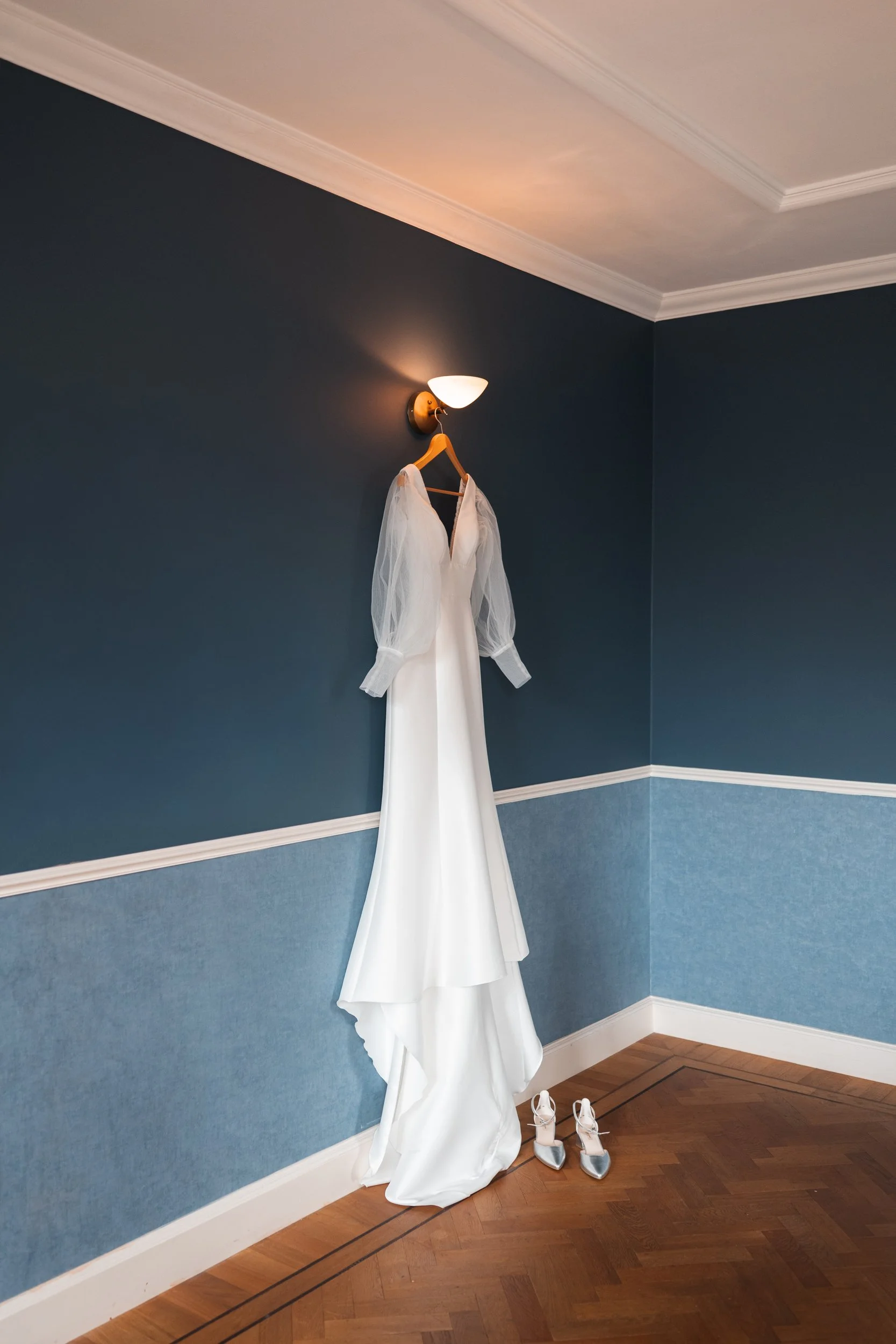 A white wedding dress with sheer long sleeves hangs from a wall-mounted light fixture in a room with blue walls. A pair of white and silver high-heeled shoes are on the wooden floor beneath the dress.