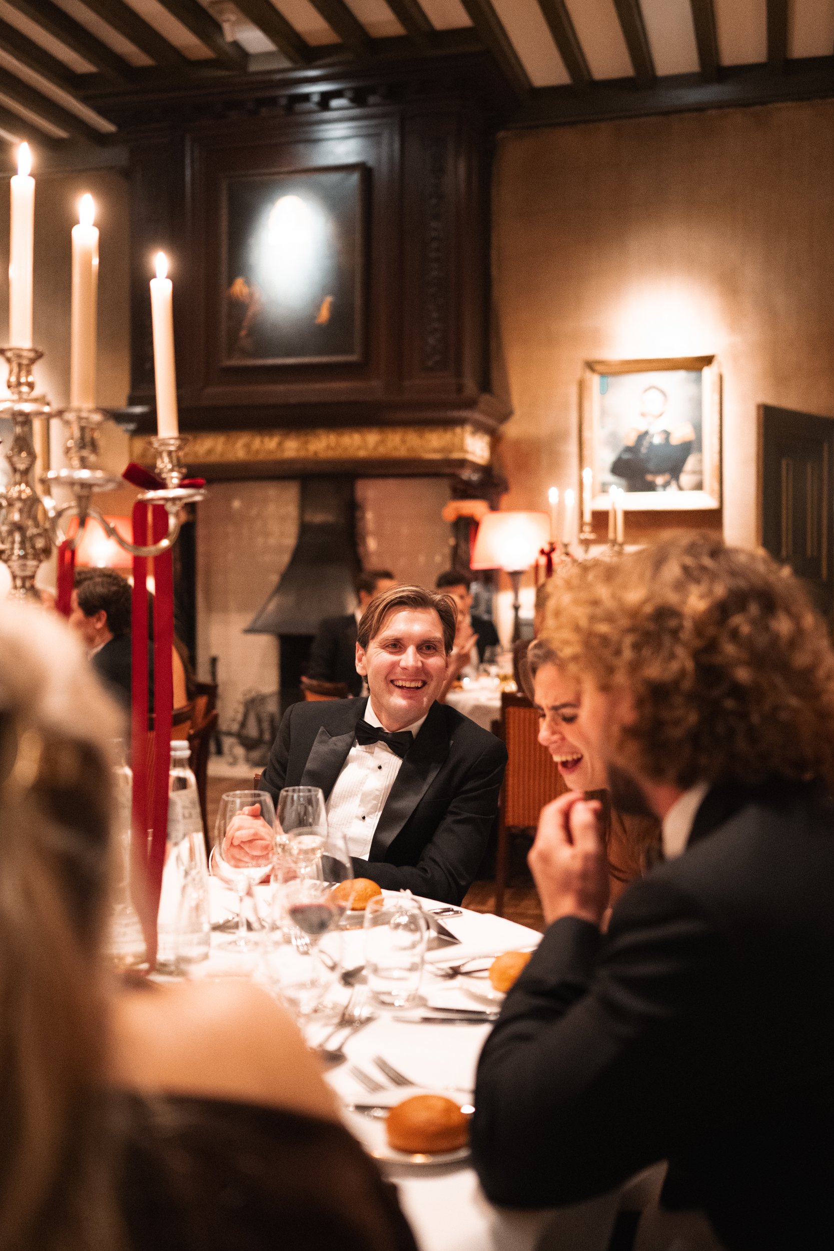 People in tuxedos laughing and talking at a dinner party in an elegant, warmly lit restaurant with candles and classic decor.