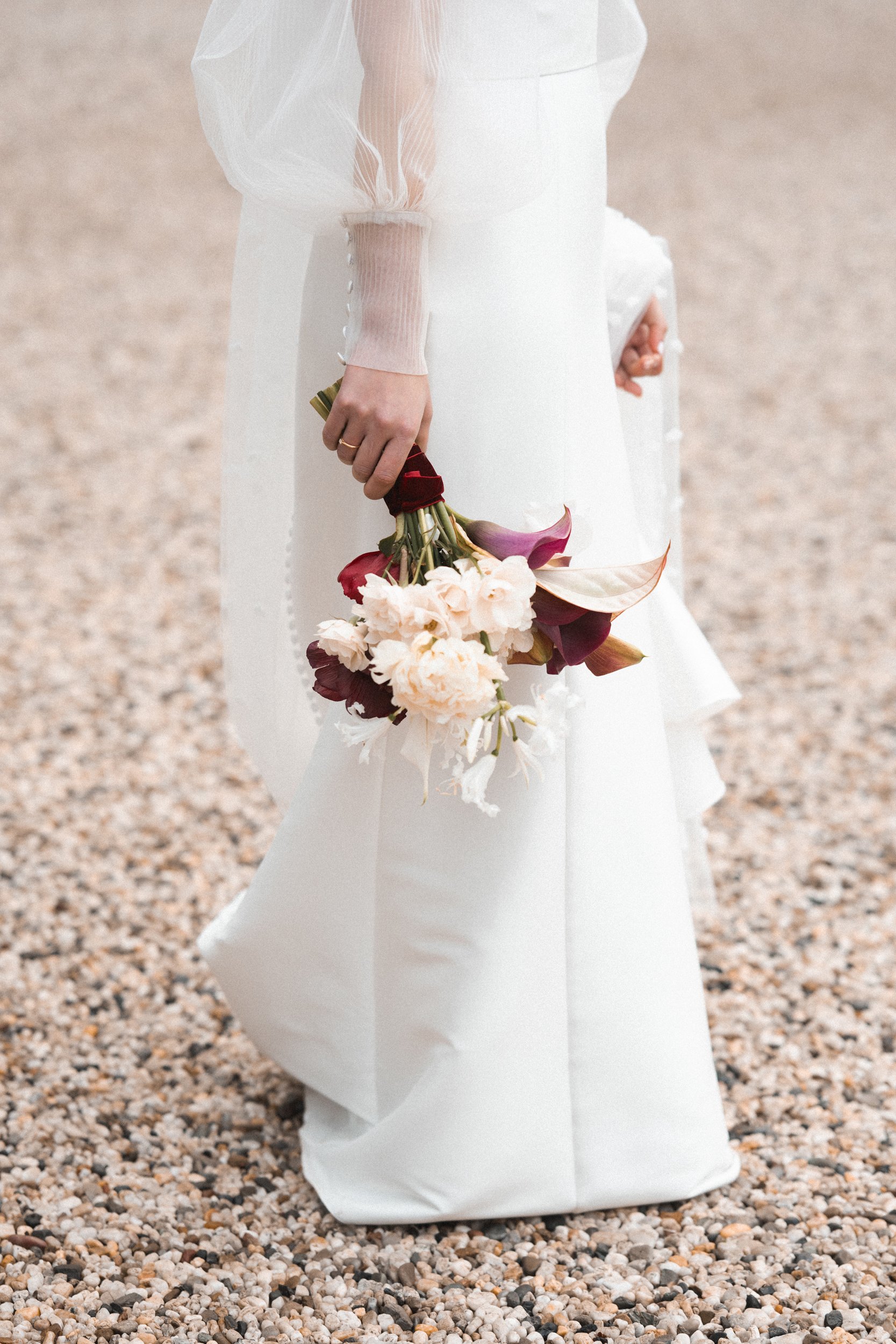 A person in a white dress holding a bouquet of assorted flowers, including lilies, roses, and peonies, standing on a gravel surface.