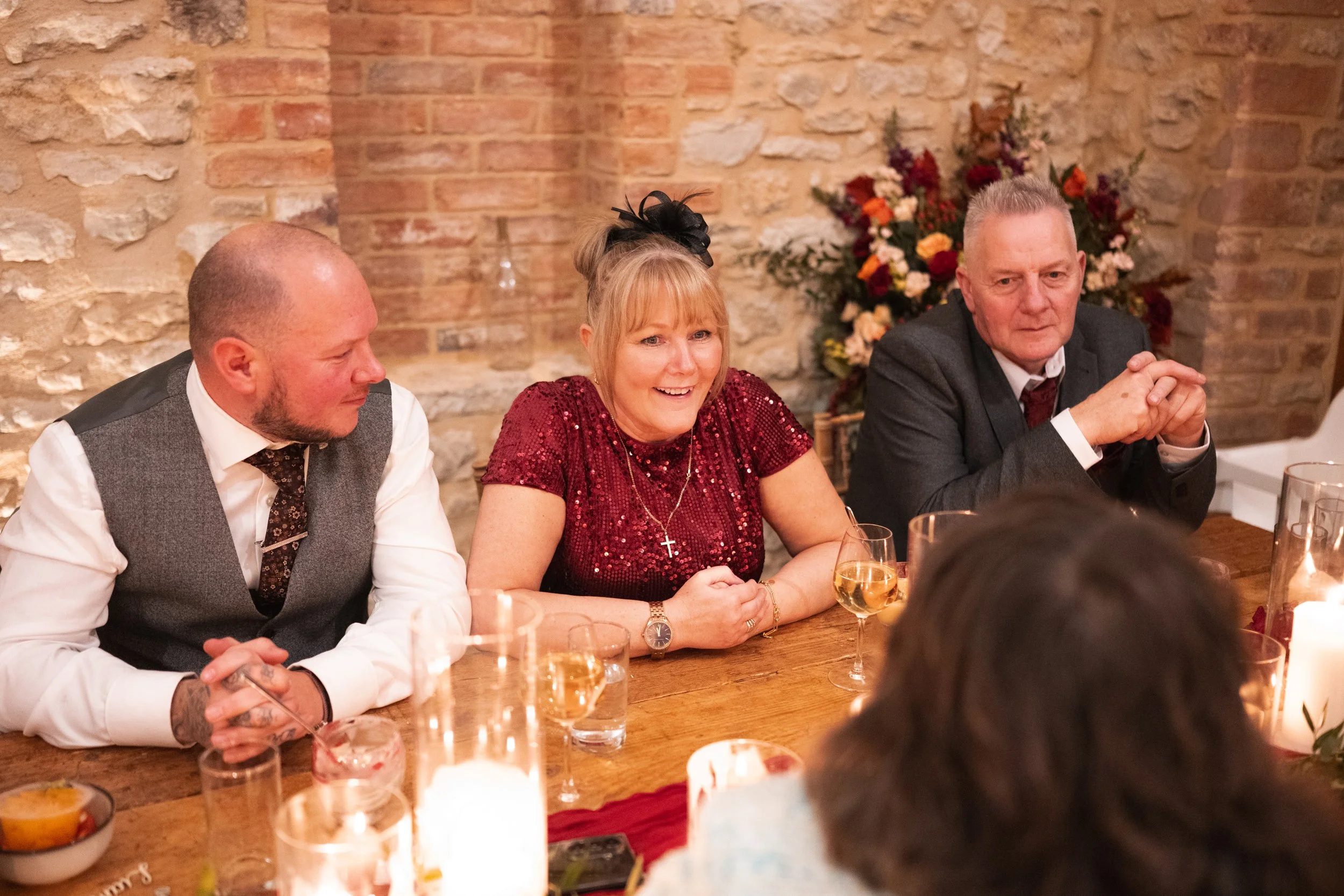 Three people sitting at a wooden table during a celebration or dinner, with drinks and a lit candle, in front of a brick wall and floral arrangement.