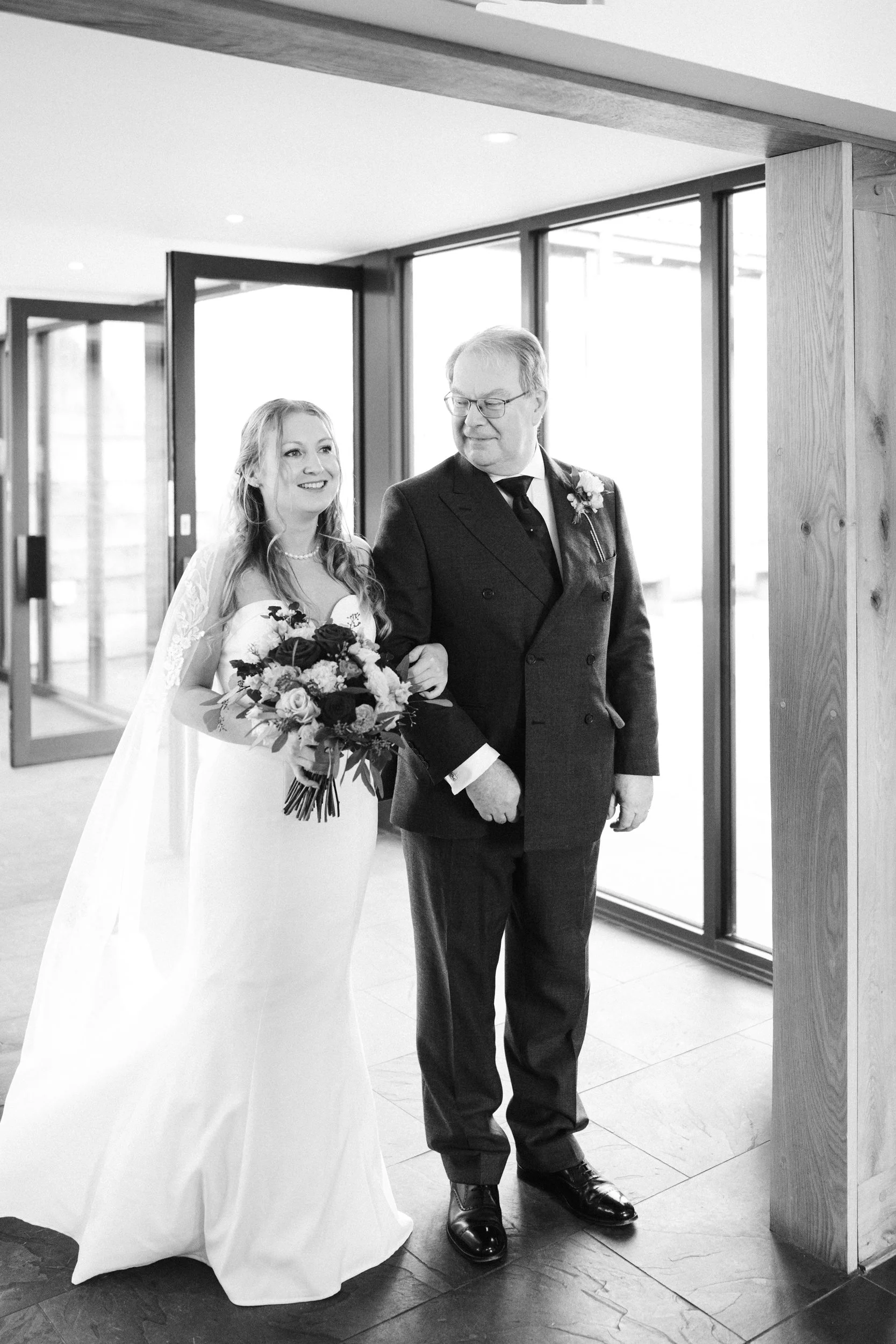 A bride walking down the aisle with a man, likely her father, holding her arm. The bride is smiling and holding a bouquet of flowers, wearing a wedding dress and veil. The man is dressed in a dark suit with a boutonniere, smiling and looking at the b