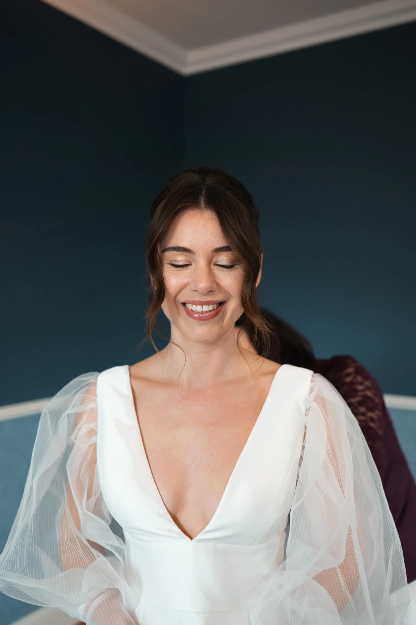A woman in a white dress smiling with eyes closed, indoors with dark blue walls.