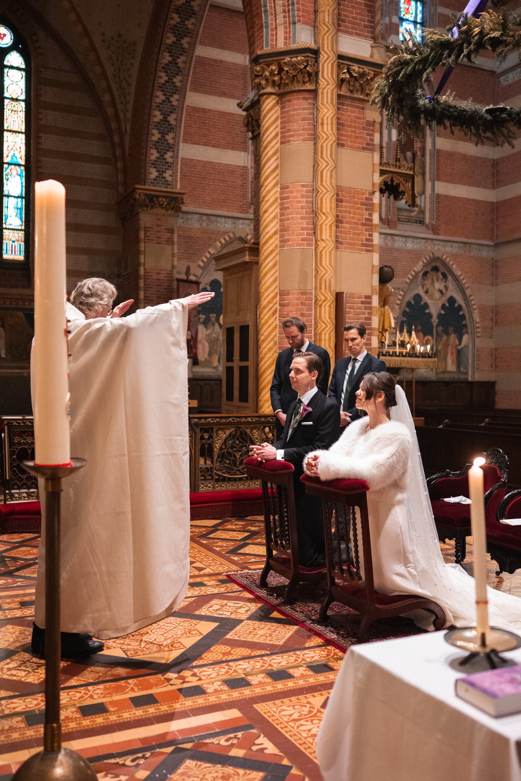 A wedding ceremony taking place inside a church with intricate patterned floors, stained glass windows, and ornate interior design. The bride and groom kneel at the altar, with the priest officiating and three groomsmen standing behind them.