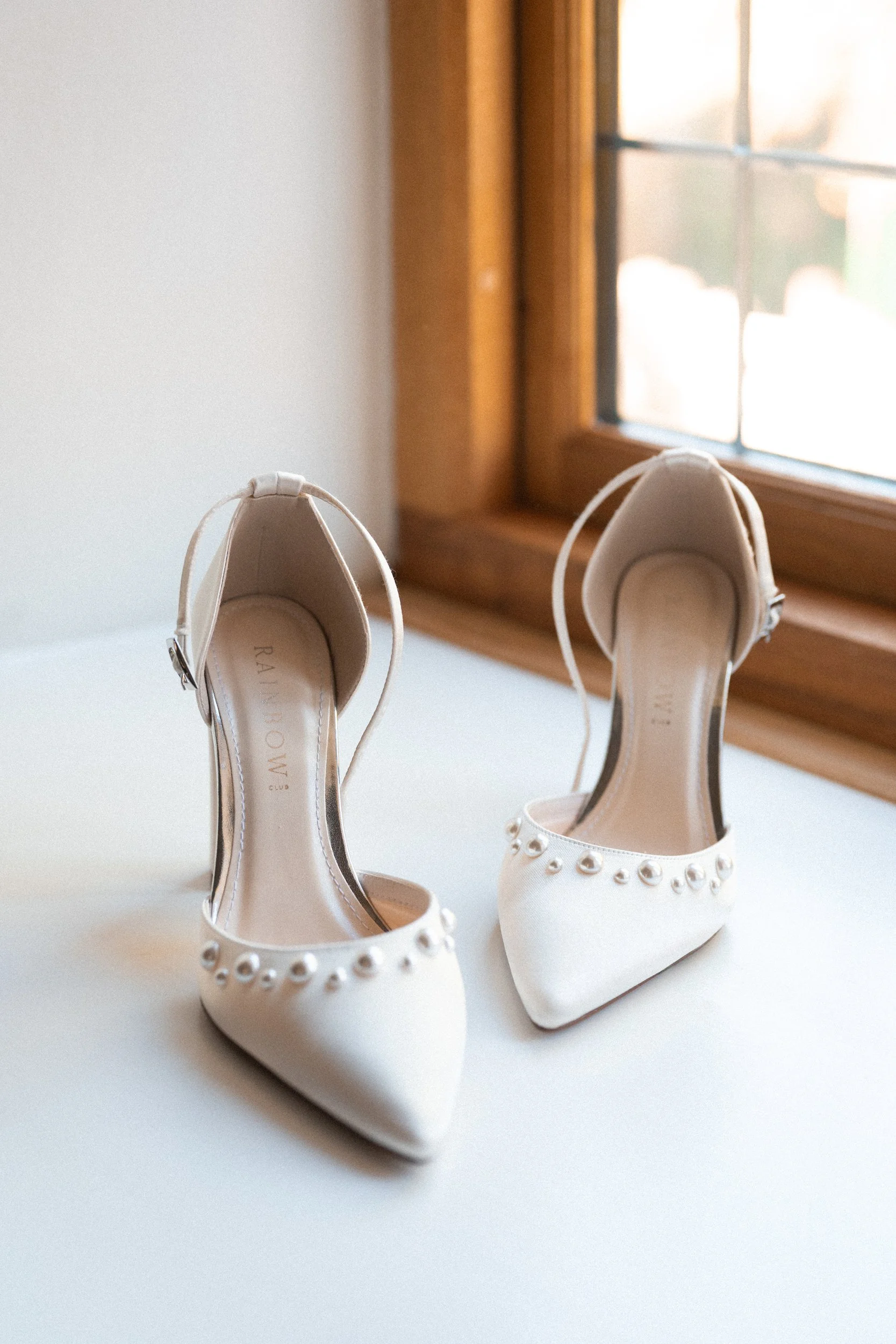 A pair of white high-heeled shoes with pearl embellishments on the toes, placed on a white surface near a window with wooden trim.