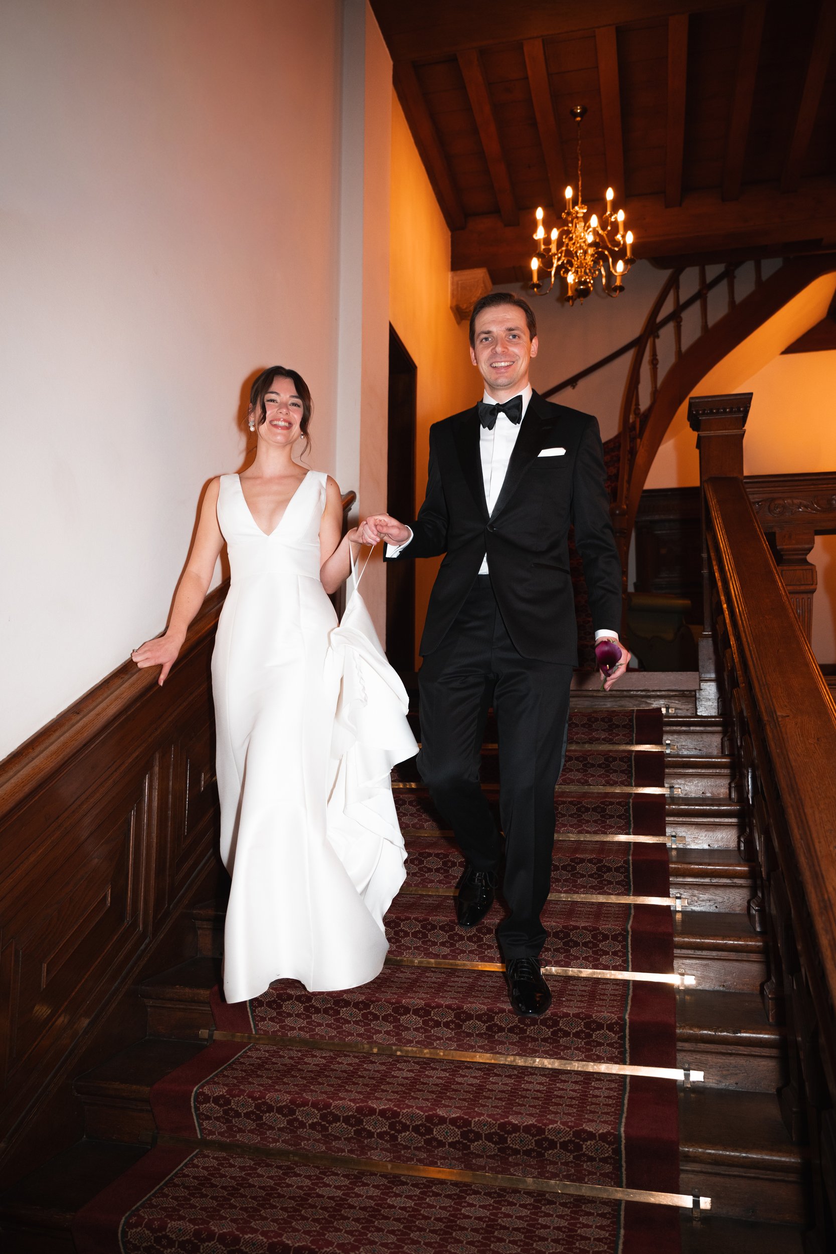 A bride in a white gown and a groom in a black tuxedo walking down a staircase at a wedding reception, holding hands and smiling.