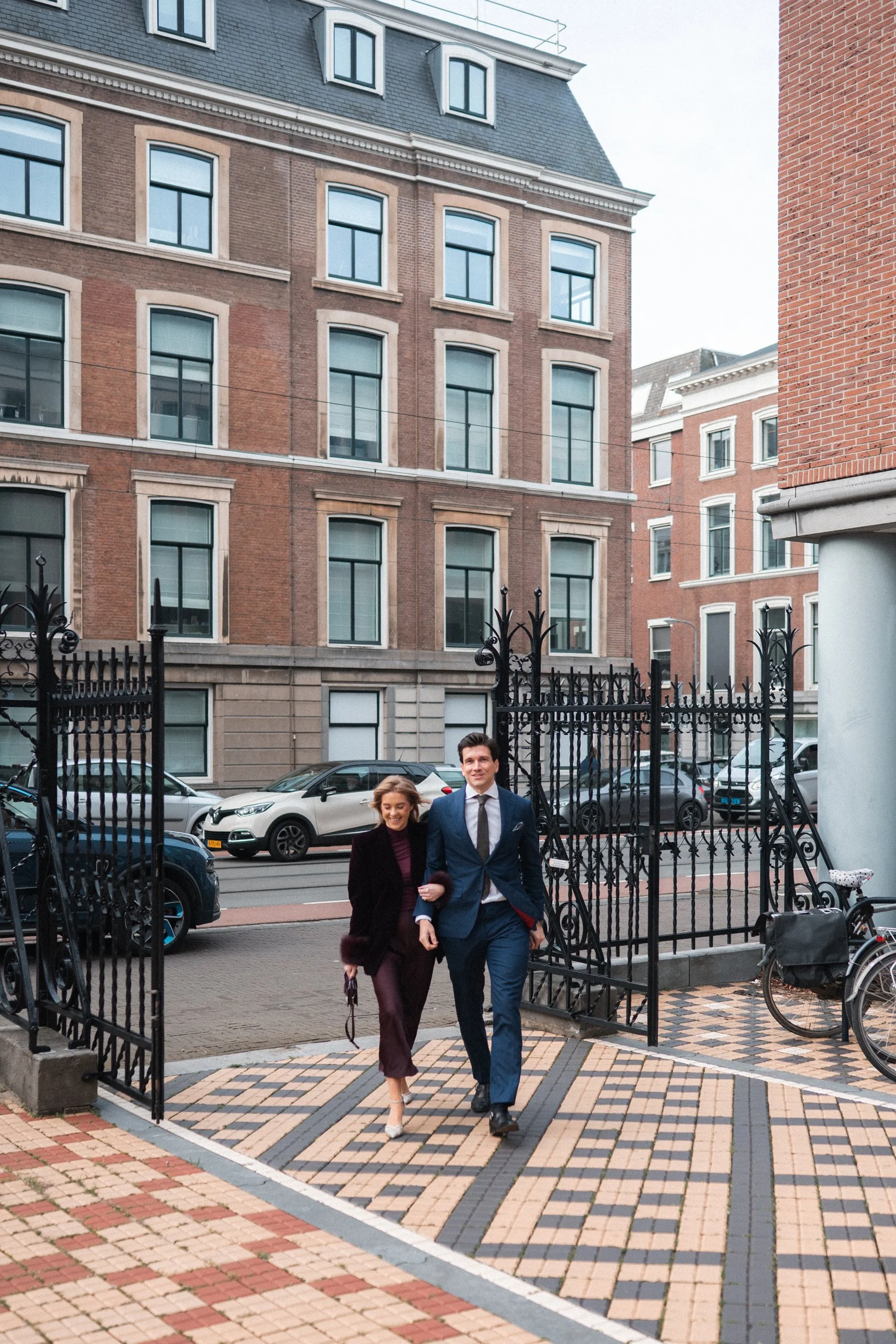 A man and a woman dressed in formal attire walking through a wrought-iron gate in an urban area, with cars parked along the street and multi-story brick buildings in the background.