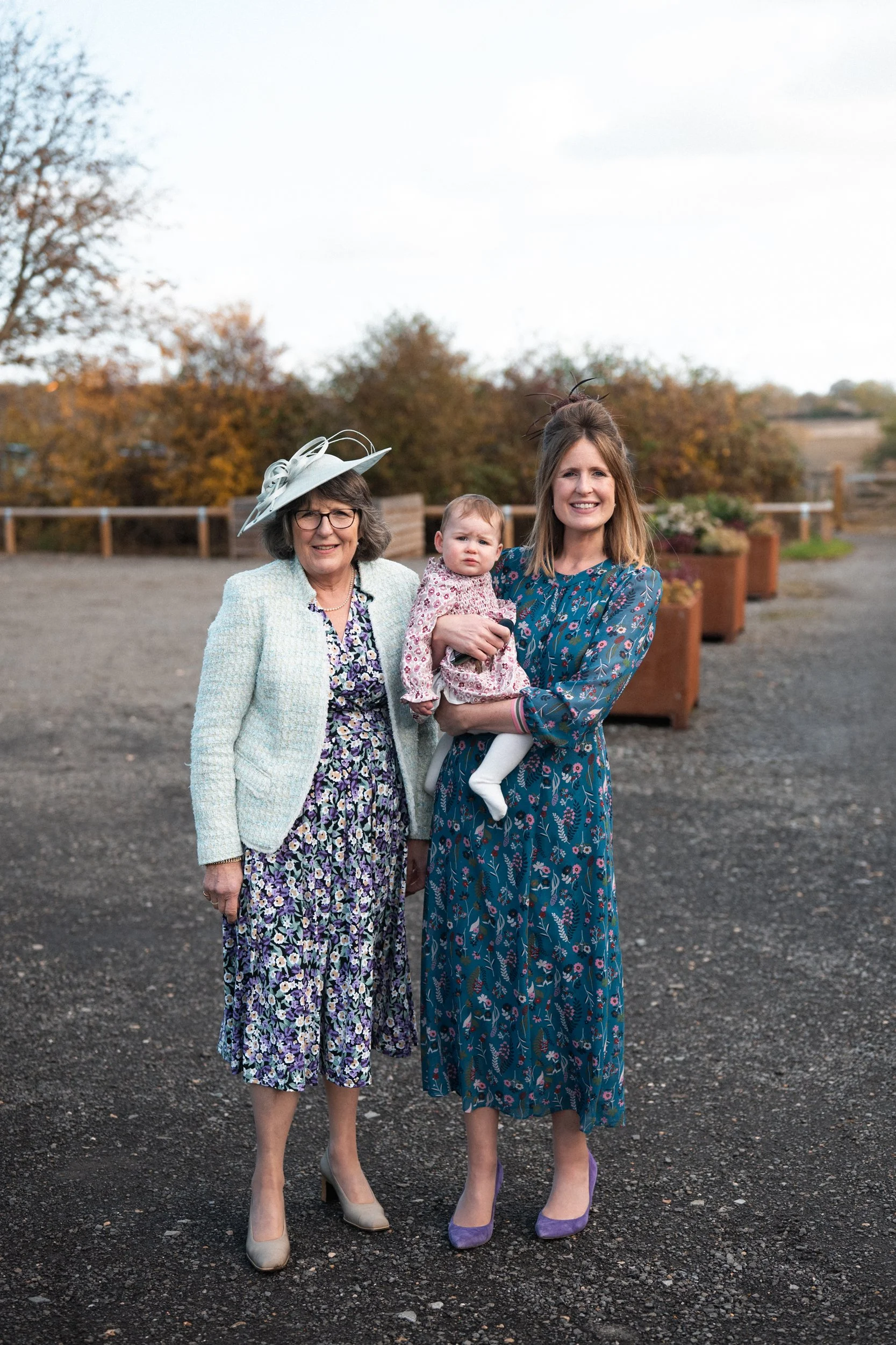 Three women and a young girl standing outdoors on a dirt path with trees in fall foliage behind them.