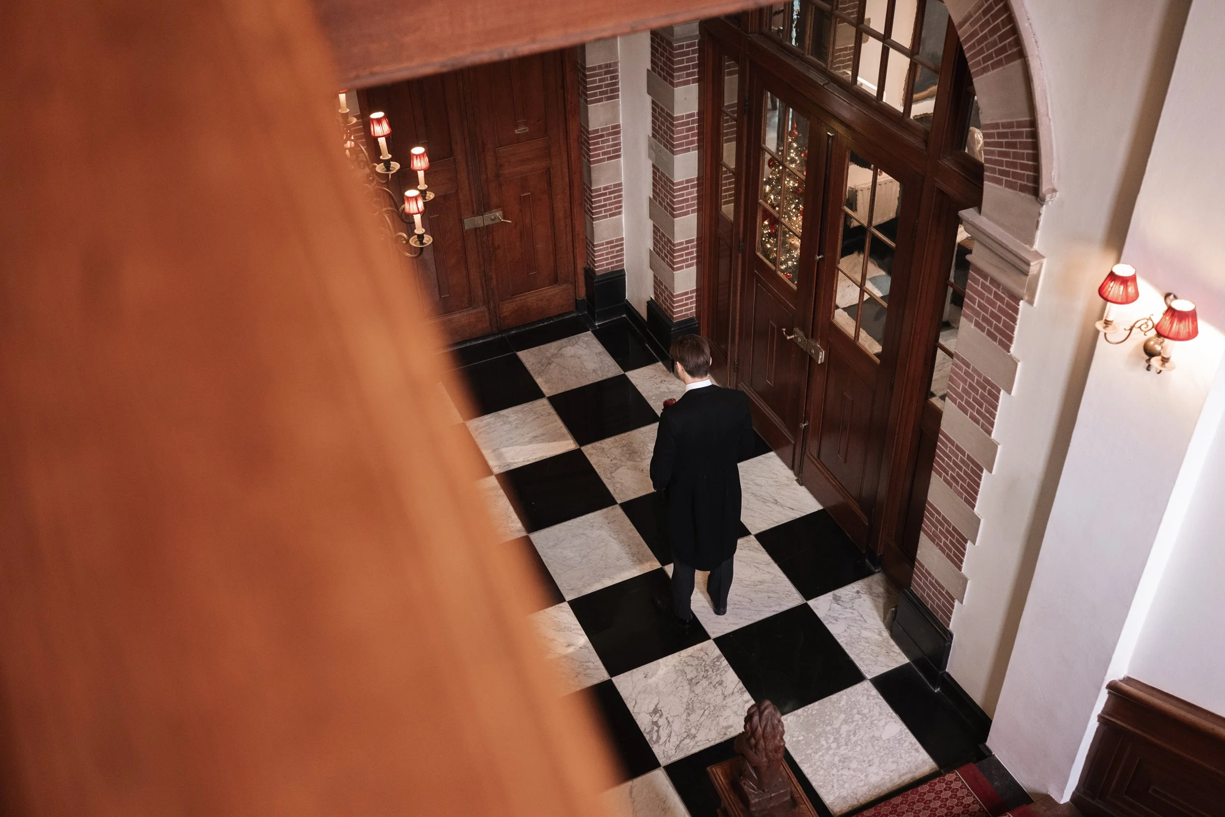 A man in formal attire walking inside a grand hotel lobby with checkered black and white marble flooring, brick walls, wooden doors, and red lamps with beige shades.
