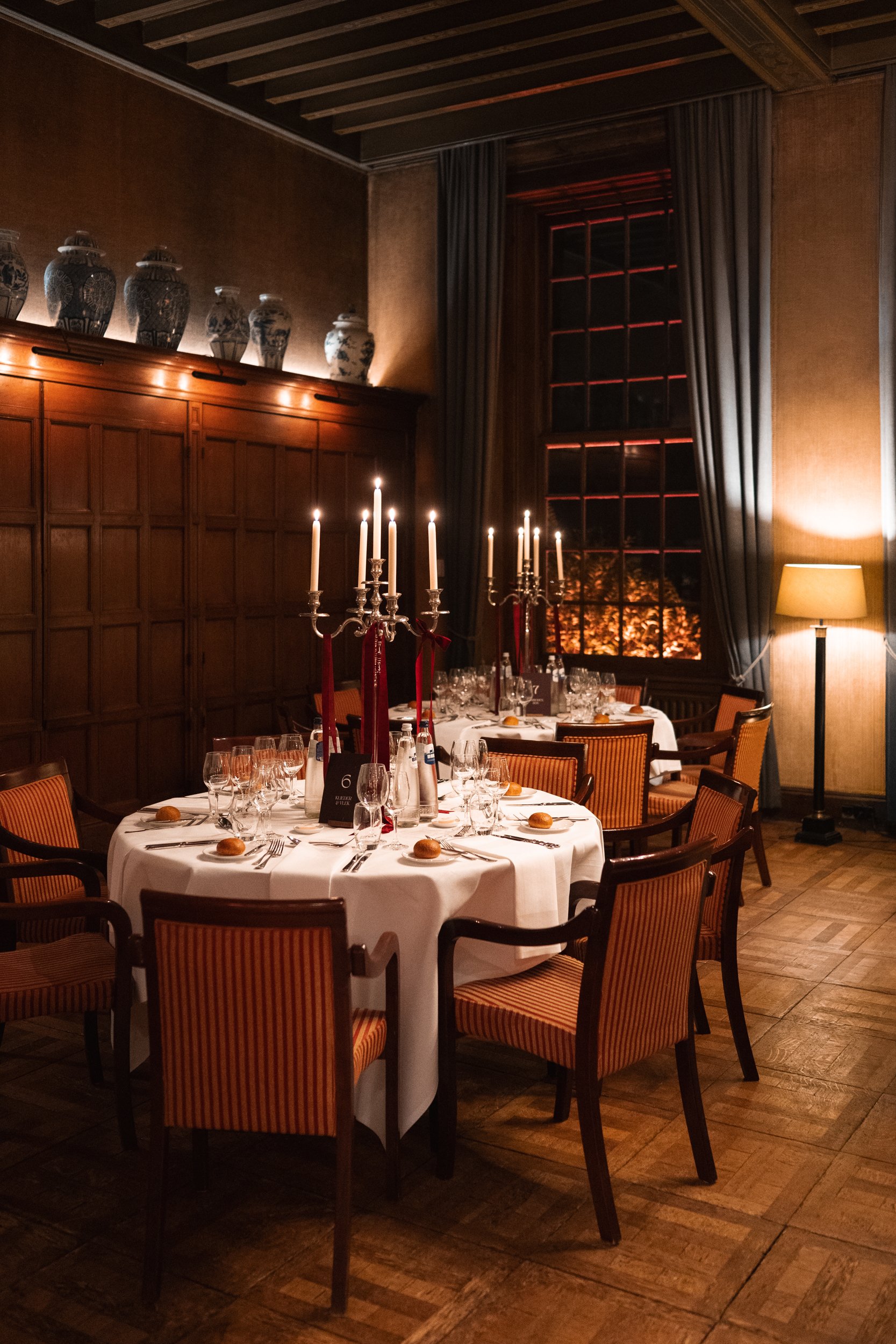 Elegant dining room with a round table set for a meal, featuring white tablecloth, multiple glasses, silverware, bread rolls, and tall candelabra with lit candles. The room has wooden paneling, a large window with curtains, decorative vases on top of