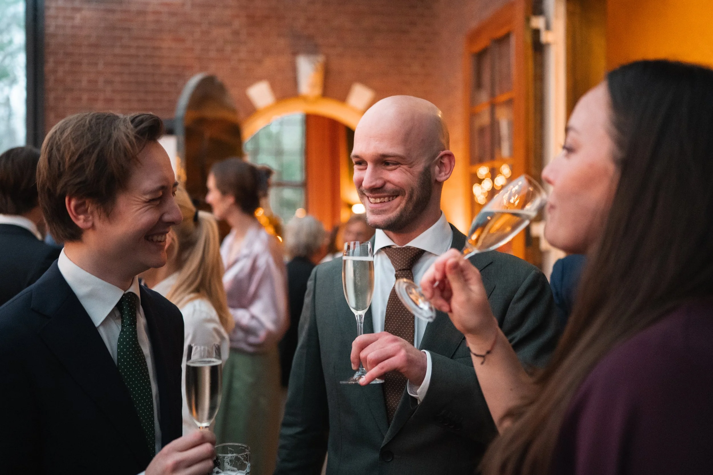 Three people in formal attire are talking and smiling at an indoor event, holding glasses of champagne.