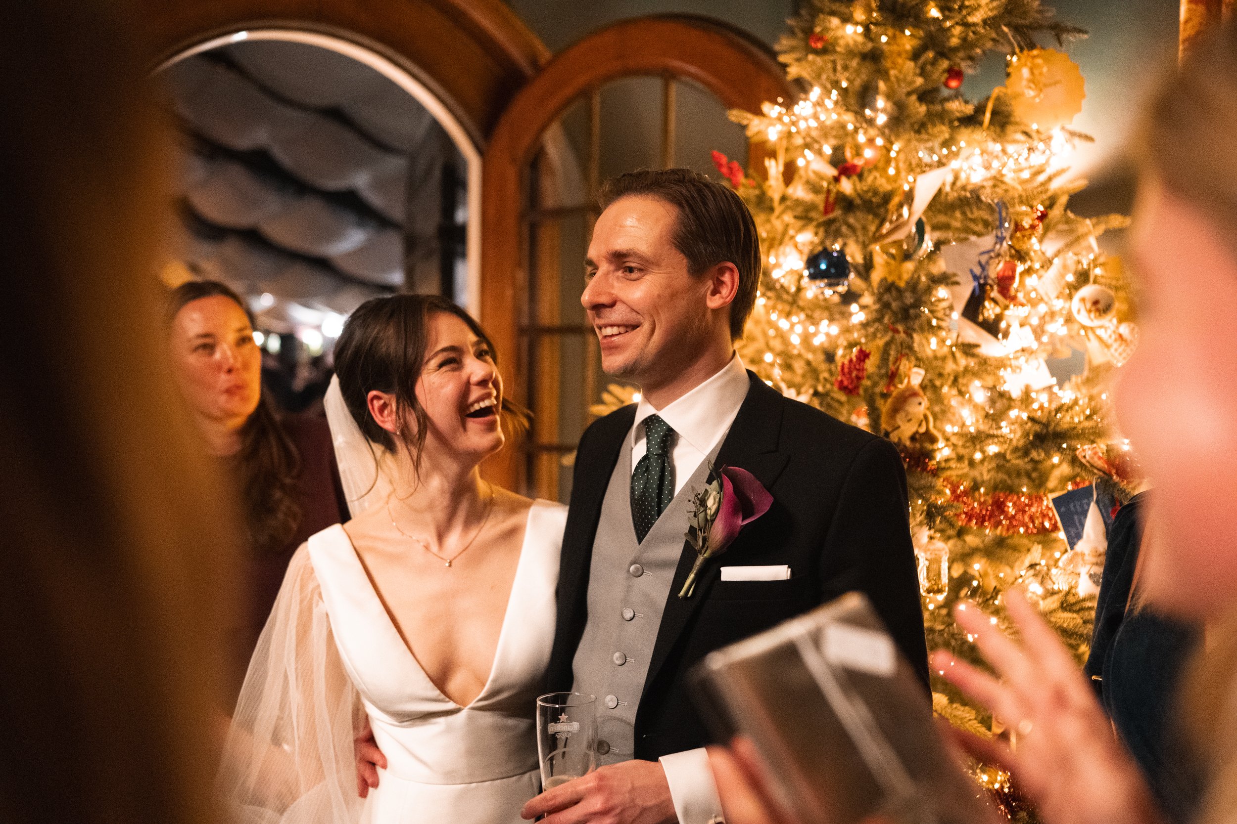 A bride and groom smiling and looking at each other at their wedding reception, standing near a decorated Christmas tree with lights and ornaments.