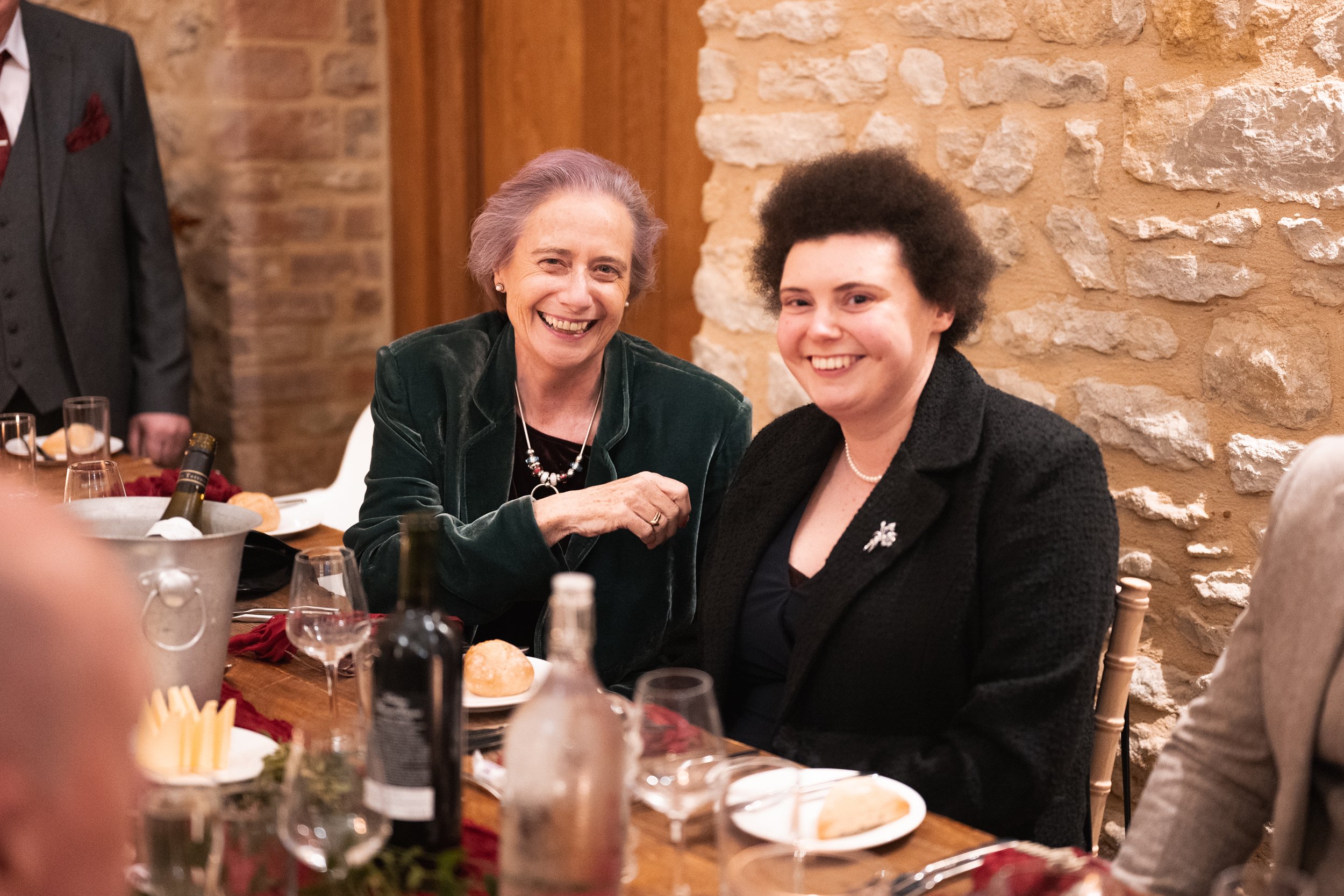Two women smiling and sitting at a dinner table during a celebration or gathering, with food and drinks on the table, and a brick wall in the background.