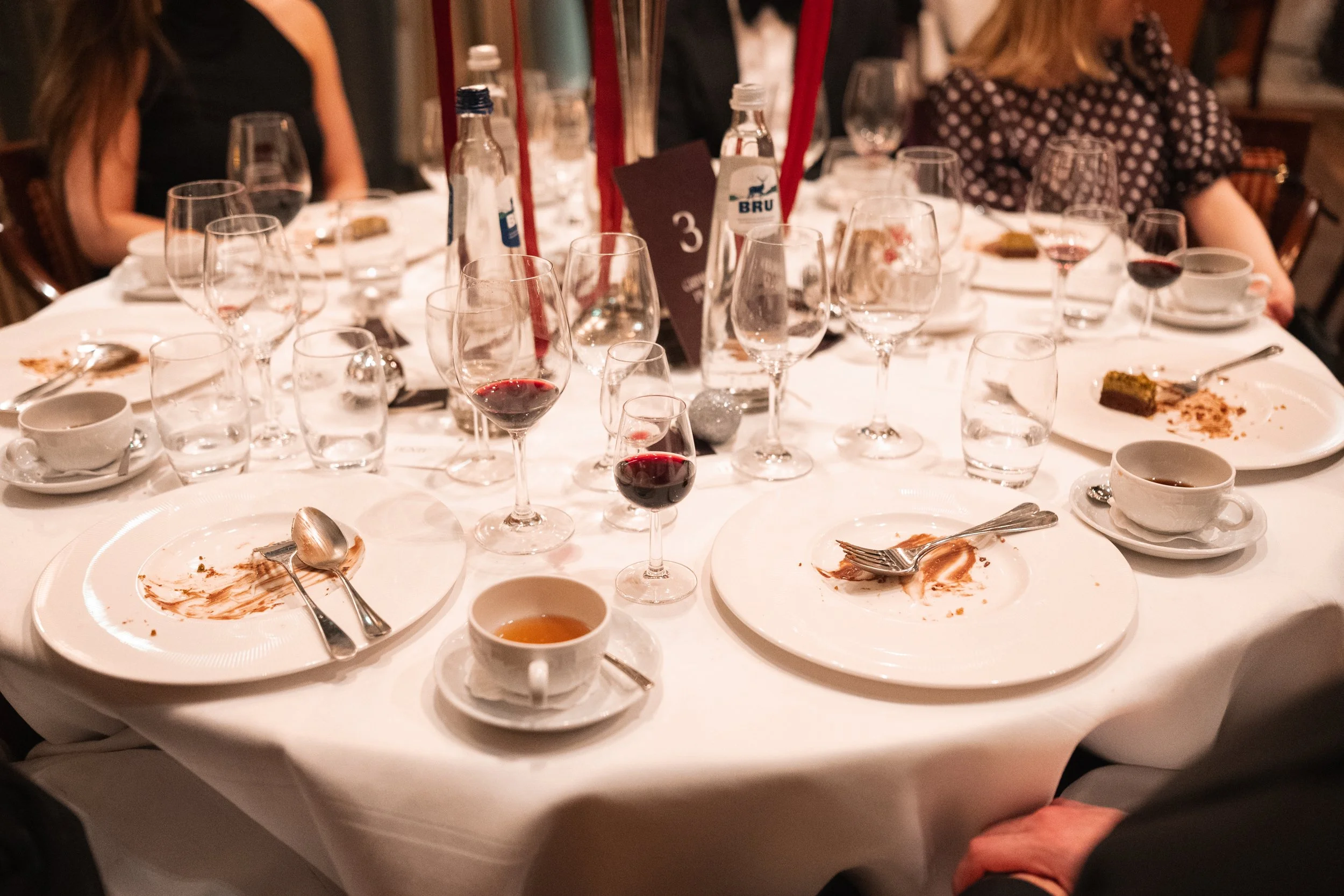 A round dining table with leftover food, empty wine glasses, water bottles, coffee cups, and silverware, with people sitting around it in a restaurant setting.