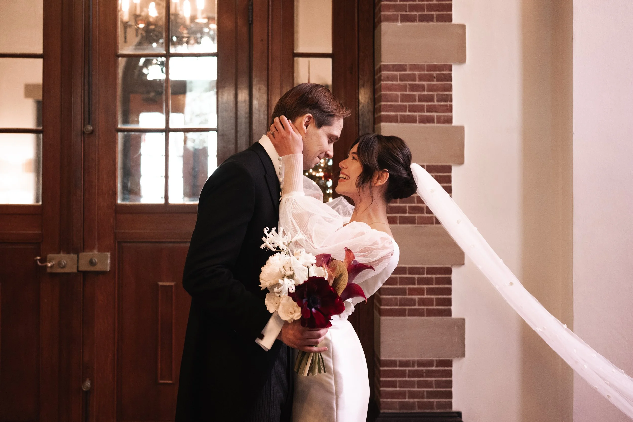 A bride and groom sharing a joyful moment indoors. The groom in a black suit holding a bouquet, and the bride in a white dress with puffed sleeves and a veil, smiling at each other.